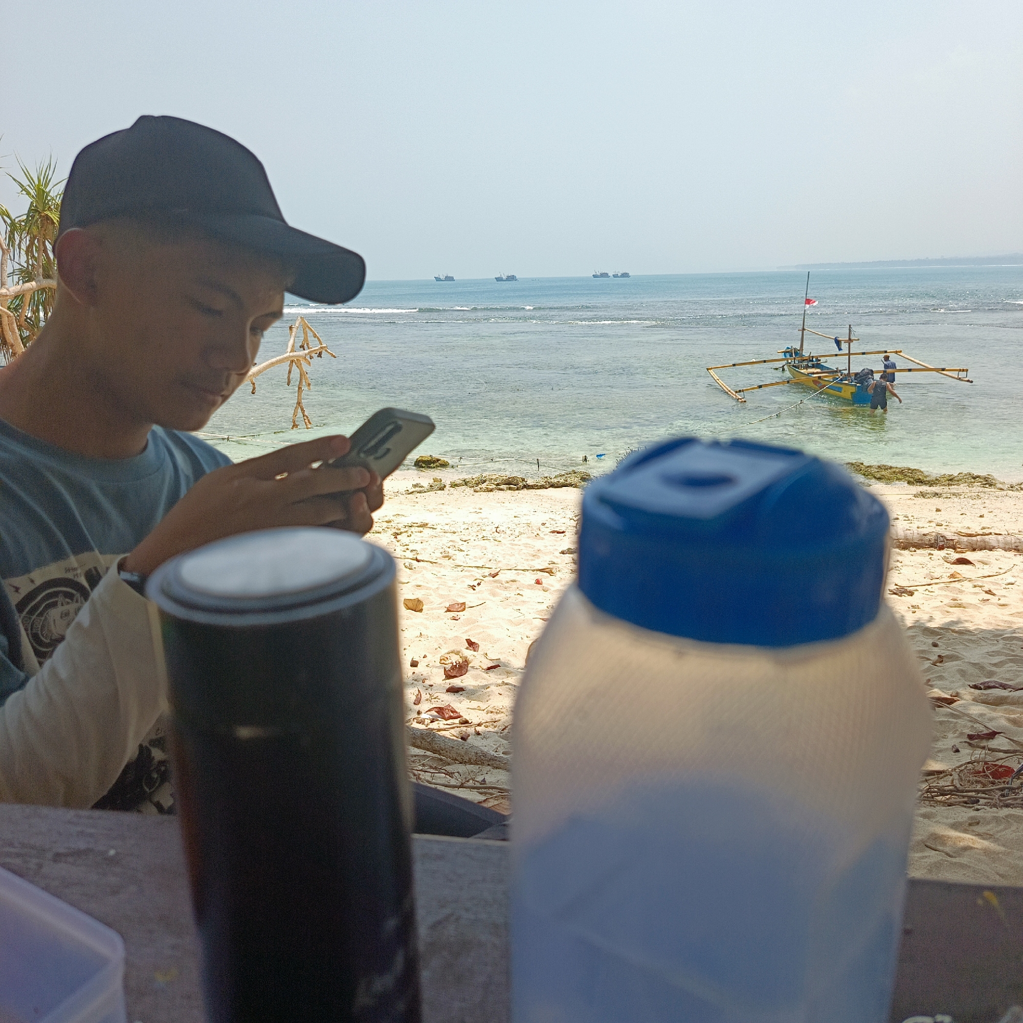 The image depicts a young person wearing a black baseball cap and a blue t-shirt, engrossed in their smartphone while sitting at a table on a sandy beach. In the foreground, two containers are visible, one a dark cylinder and another a white plastic container with a blue lid. Beyond the table, the beach stretches towards the ocean. The sea is calm with a few boats anchored offshore, and in the distance, several cargo ships are visible on the horizon. The sky is clear and bright, suggesting a sunny day. The location is identified as Pengubaian, Indonesia, a coastal area known for its beaches and maritime activities.