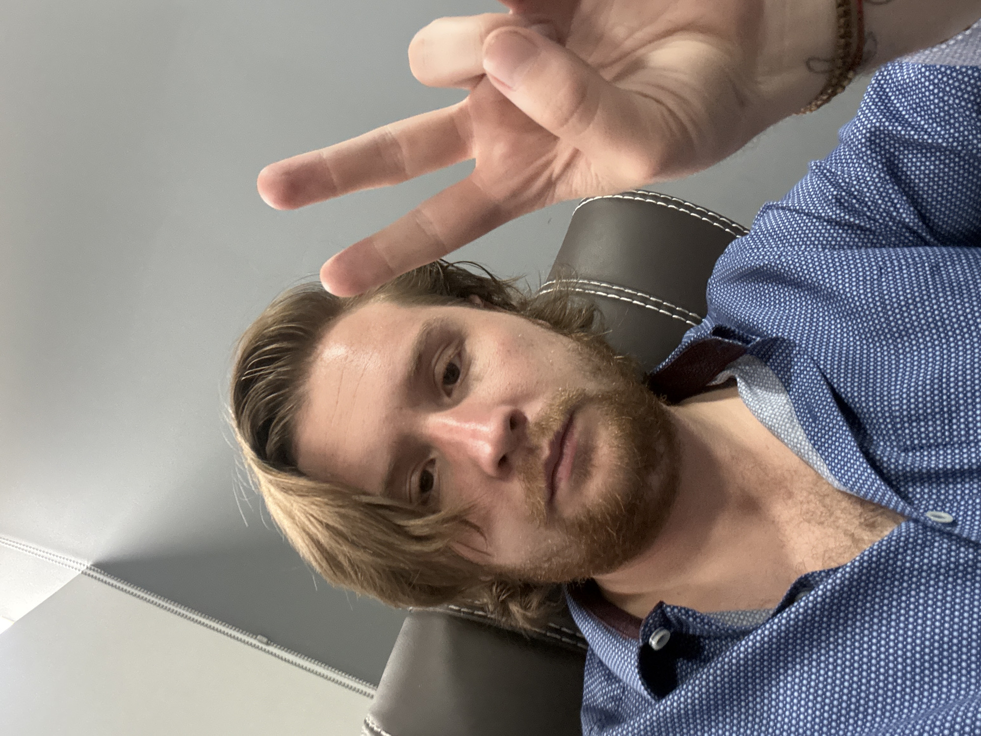 A selfie taken by a light-skinned male with brown hair and a red beard. He is wearing a blue patterned shirt and sitting in a black leather chair. His right hand is raised, displaying a "peace" sign. A bracelet is visible on his right wrist. The background includes a gray ceiling and wall. There is no information to suggest the location is in the city of Cuautitlán Izcalli, Mexico.