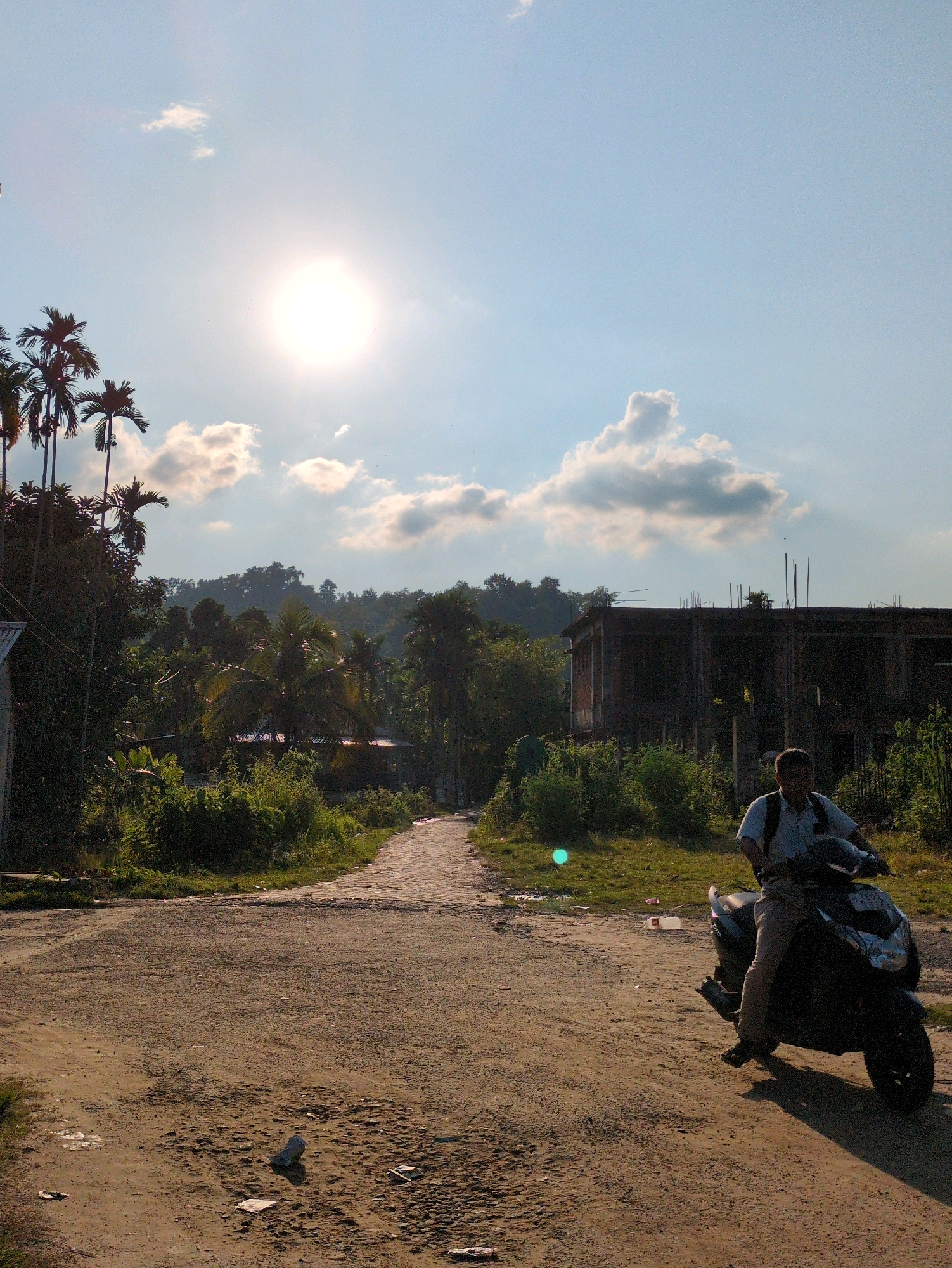 A man is riding a dark-colored scooter down a dirt road in Borchora, India. He is wearing a light blue collared shirt and tan pants, with a dark backpack on his shoulders. The sun is high in the sky, casting long shadows. To the left of the road, there are several palm trees and dense foliage. In the background, there is a partially constructed building with exposed rebar. The road is uneven and dusty, with some scattered debris. The sky is mostly clear with a few scattered white clouds.