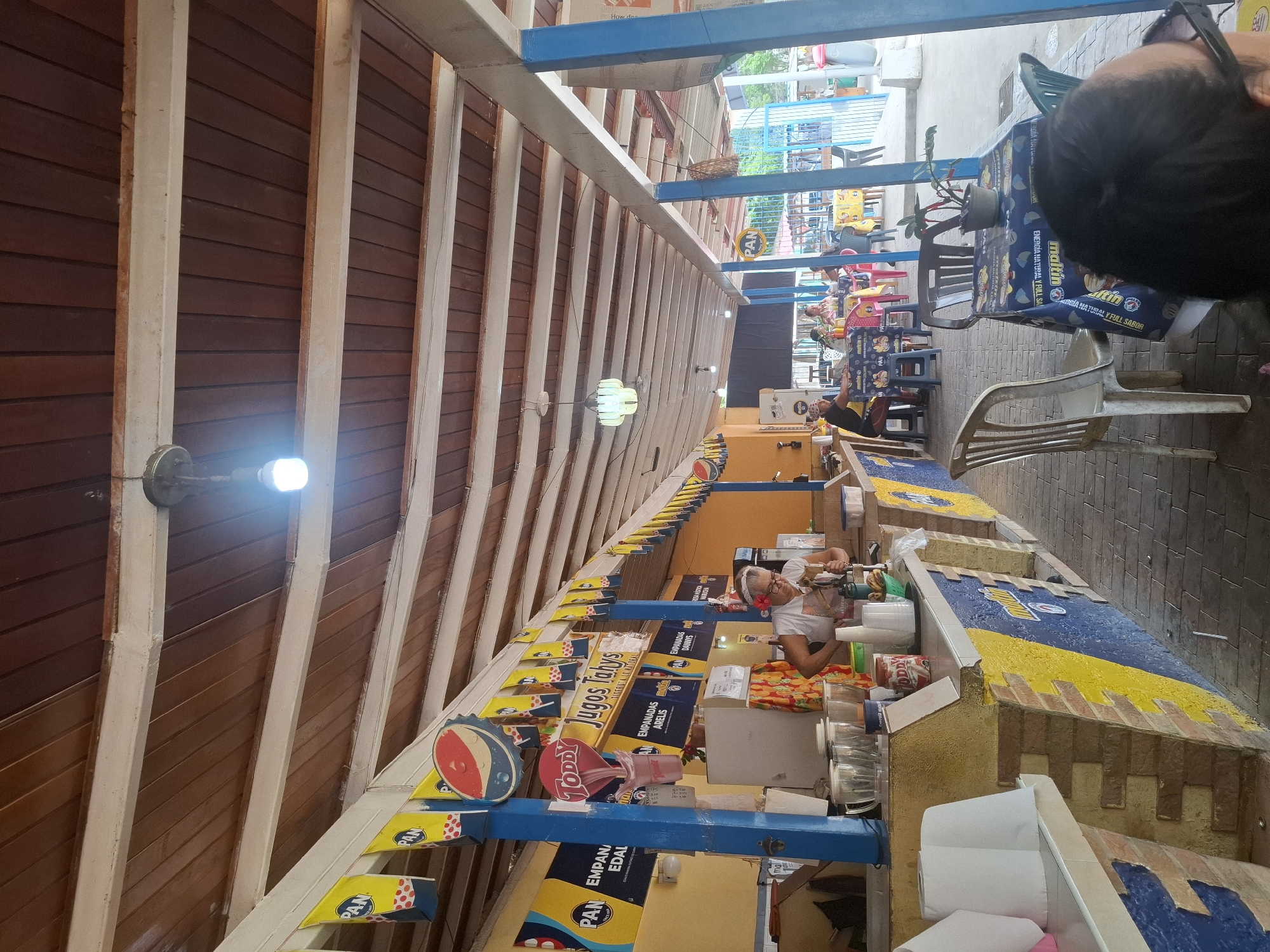 The image captures a vibrant open-air market or food court in El Valle del Espíritu Santo, Venezuela. The perspective is taken from a slightly elevated point, looking down a long, covered walkway lined with food stalls and dining areas.
A prominent feature is the overhead wooden ceiling or awning, composed of dark wooden planks separated by lighter beams, with bright artificial lights illuminating the space. Along the right side of the frame, several food stalls extend into the background. These stalls have counters decorated with blue and yellow designs, some mimicking brickwork, and are adorned with various commercial banners and signs.
Multiple people are present, indicating a busy atmosphere. In the mid-ground, a vendor, possibly an older woman with glasses, is actively engaged behind a counter, appearing to prepare food or drinks using blenders and other equipment. Further down, patrons are seated at tables covered with branded blue tablecloths, enjoying their meals or waiting for service. The top of a person's head, wearing glasses, is visible in the top right foreground, looking towards the market activity.
The scene conveys a casual dining and shopping experience. Visible text on banners and signs includes "PAN" (repeatedly), "TODDY," "Jugos Tahys," "EMPANADAS ARELIS," "EMPANADAS DANNYS," and "EMPANADA EDAL." The brand "maltin" is frequently displayed, often alongside the phrase "ENERGIA NATURAL Y FULL SABOR." Some prices are also legible on small display signs. The flooring throughout the market appears to be made of paved stones or tiles. The setting suggests daytime, despite the use of artificial lighting, contributing to a lively and bustling environment.