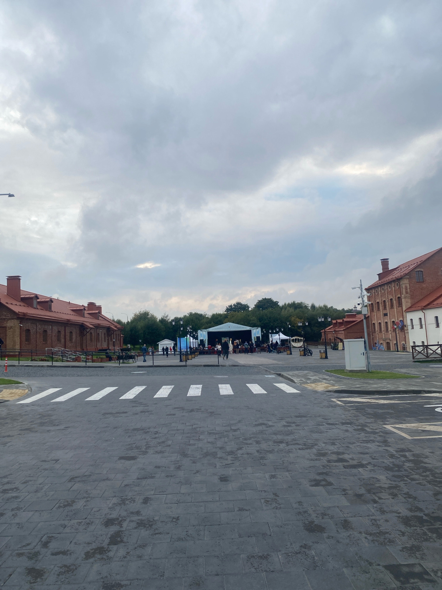 This is a photograph of an outdoor scene in Minsk, Belarus, likely at the "Belaruskaya Vyalikaya" museum complex in the Pervomaysky district. The image shows a wide, paved area with a pedestrian crossing in the foreground. To the left, there is a long, red-brick building with a red roof, characteristic of the museum's historical architecture. On the right, another brick building with a red roof is visible, featuring arched windows.
In the middle ground, there is an open space with a stage or performance area set up under a large, white tent-like structure. Several people are gathered around the stage, some seated. There are also food stalls or market booths with tents and tables set up, and a horse-drawn carriage. Trees and greenery form a backdrop behind these structures. The sky is overcast with light gray clouds. The overall impression is of a public event or gathering taking place in a historical urban setting.