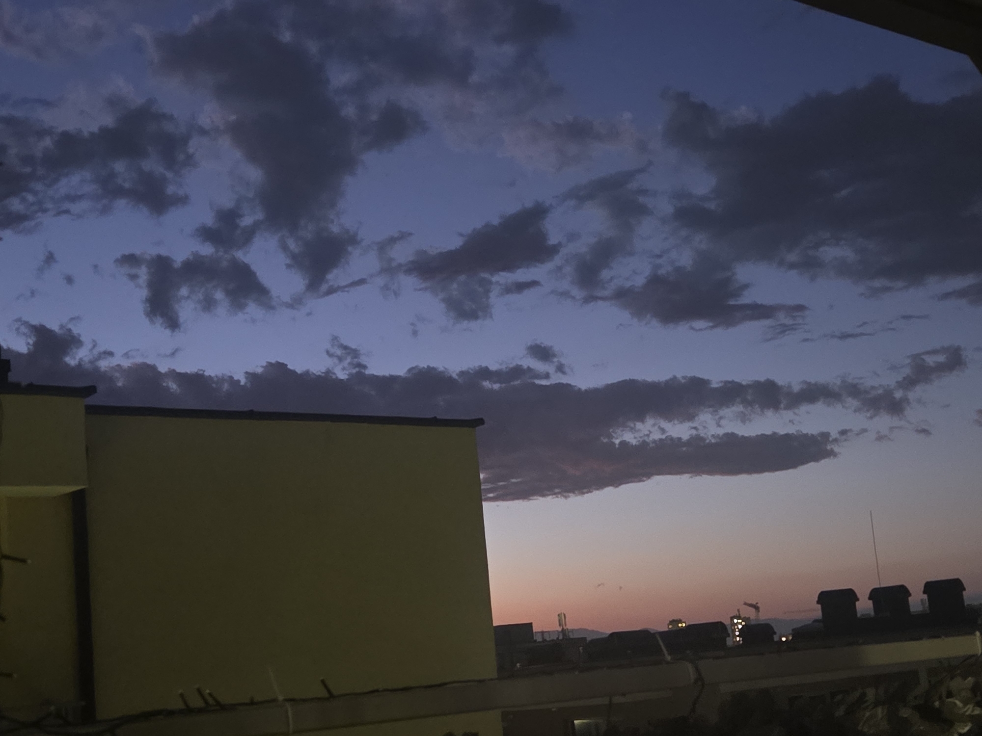The image shows a twilight scene, likely taken from a high vantage point in an urban setting. In the foreground is a portion of a building with a yellow facade. Behind this building, a cityscape with various rooftops, vents, and distant structures is visible against a sky exhibiting a gradient of colors from deep blues to pinks and oranges. The sky is filled with dark, ominous clouds. The location is unknown, as well as city and country.