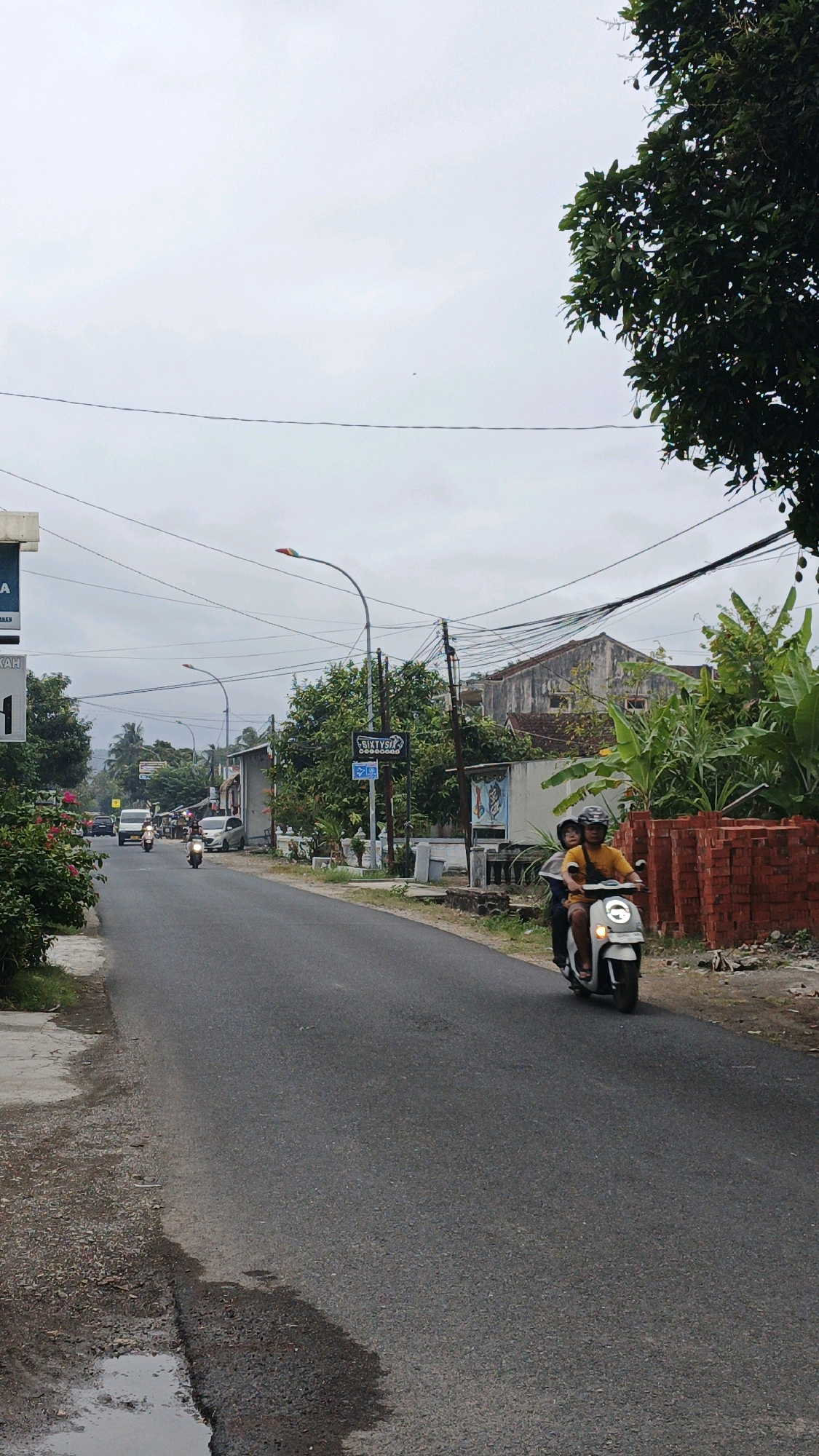 The image captures a view of a street in Parigi, Indonesia, under an overcast sky. A paved road runs through the center of the frame, with a motorcycle carrying two individuals riding away from the camera on the right side of the road. The rider wears a yellow shirt, and the passenger wears a gray shirt. Buildings and lush green vegetation line the sides of the road, with electrical wires strung overhead. A street sign with "SIKTYSTA" is visible on the right side of the road, near a lamppost with a multi-colored light. Piles of red bricks are stacked on the right verge. The left side of the road features a sidewalk and more greenery, with a puddle of water visible on the pavement near the bottom left corner. Several other vehicles, including cars and motorcycles, are present further down the road, suggesting moderate traffic. The overall scene depicts a typical day in a suburban or semi-rural area of the city.