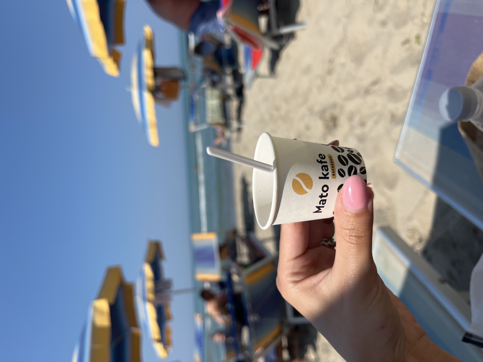 A person's hand, with pink painted fingernails and a ring on the ring finger, holds a small white paper cup with the logo "Mato kafe EXPRESSO" printed on it. A white stirring stick protrudes from the cup. The cup is positioned in the foreground, with a blurred background of a sandy beach. Beach umbrellas with yellow and blue stripes are visible, along with beach chairs and people relaxing. The ocean and a clear blue sky are in the distance. The scene appears to be a sunny day at a beach in Bashkia Durrës, Albania.