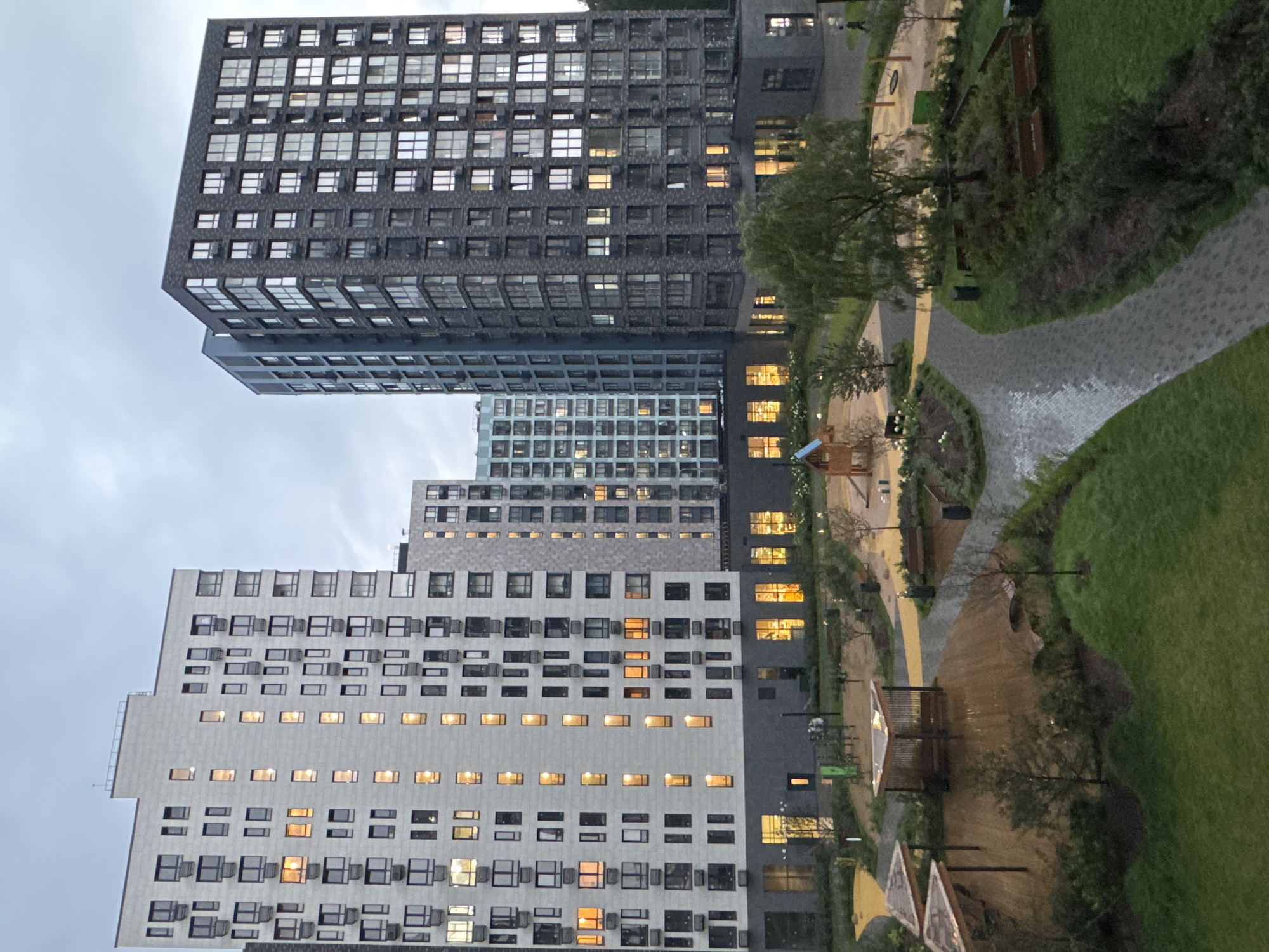 The image depicts a modern residential complex in the city of Kommunarka, Russia, on a cloudy day. The foreground features a neatly landscaped courtyard with a paved walkway, grassy areas, and a wooden deck with sheltered seating areas. A children's playground with wooden structures and swings is visible in the mid-ground. In the background, imposing multi-story apartment buildings dominate the skyline. The buildings are characterized by their uniform, geometric design, with numerous windows and balconies. Some windows are illuminated, suggesting occupancy. The overall atmosphere is one of contemporary urban living.