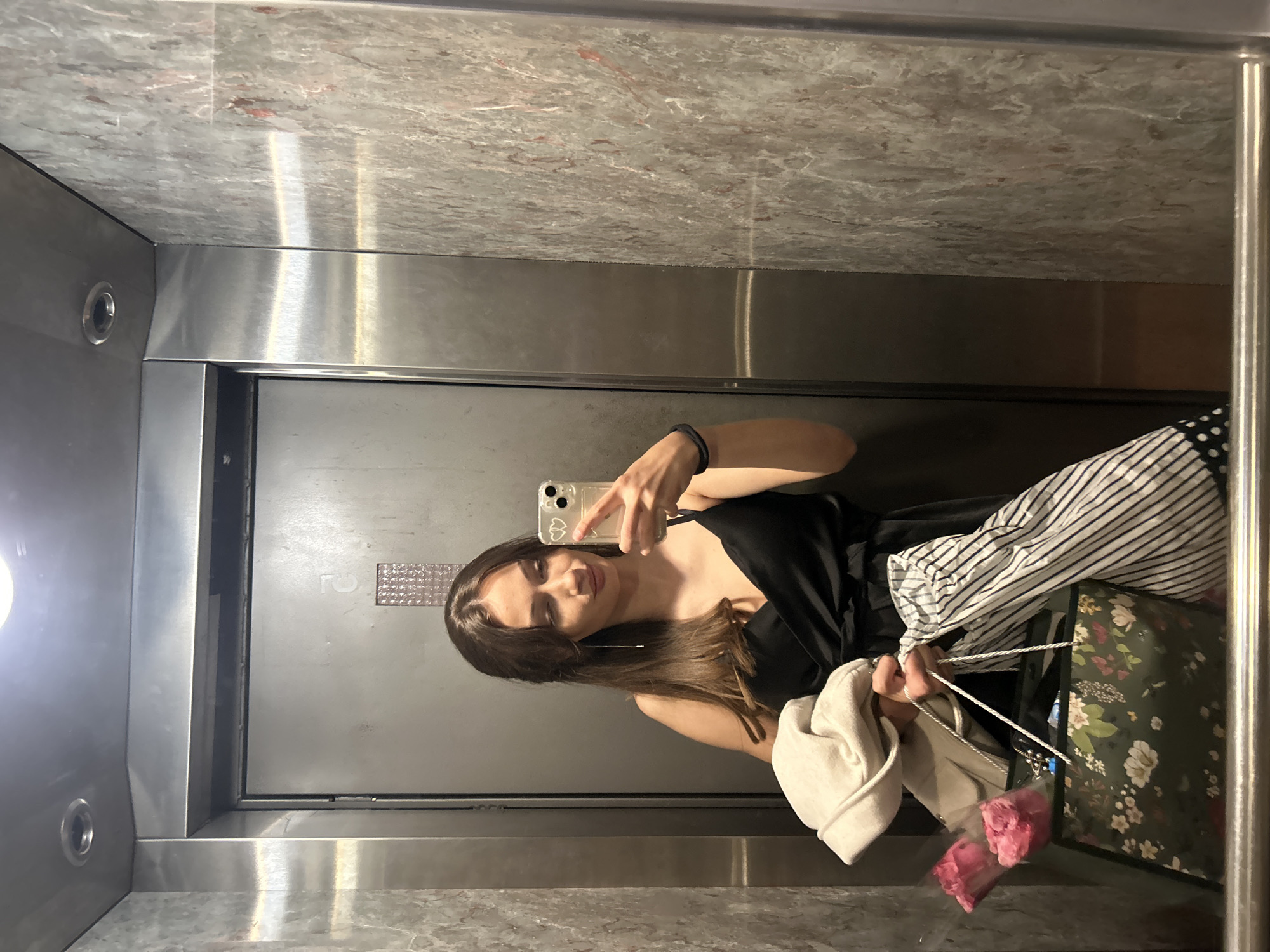 A woman with long brown hair is taking a selfie in an elevator. She is wearing a black top and striped pants. She is holding shopping bags and a white jacket. The elevator has a stainless steel interior with marble accents. The door has a number '2' on it. It is not possible to identify the location within the city of Burgas, Bulgaria, from this image alone.