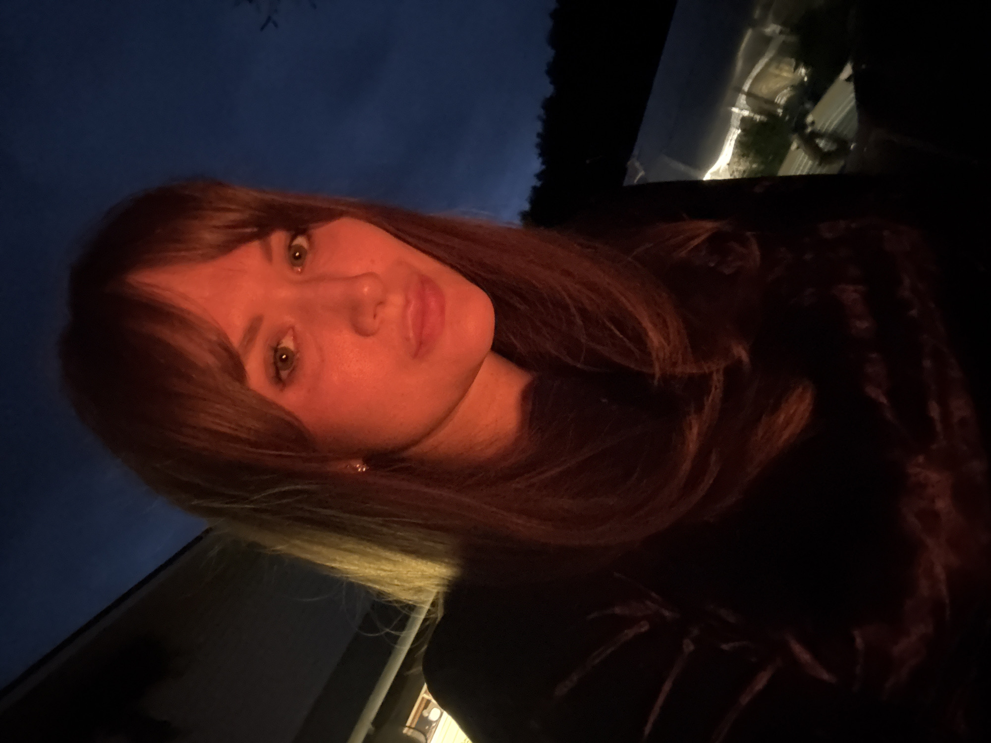 A woman with long, auburn hair and bangs poses for a selfie in the dimly lit evening. Her face is illuminated by a warm, orange light, likely from a nearby fire. She is wearing a dark-colored, possibly velvet, top. In the background, the dark sky transitions to a deeper blue, with the silhouette of trees visible against the horizon. To the right, a structure with lights on is partially visible, suggesting an urban or suburban setting. The scene evokes a cozy, possibly outdoor gathering during twilight. The exact location in Chaikovsky, Russia, cannot be determined from the visual information alone.