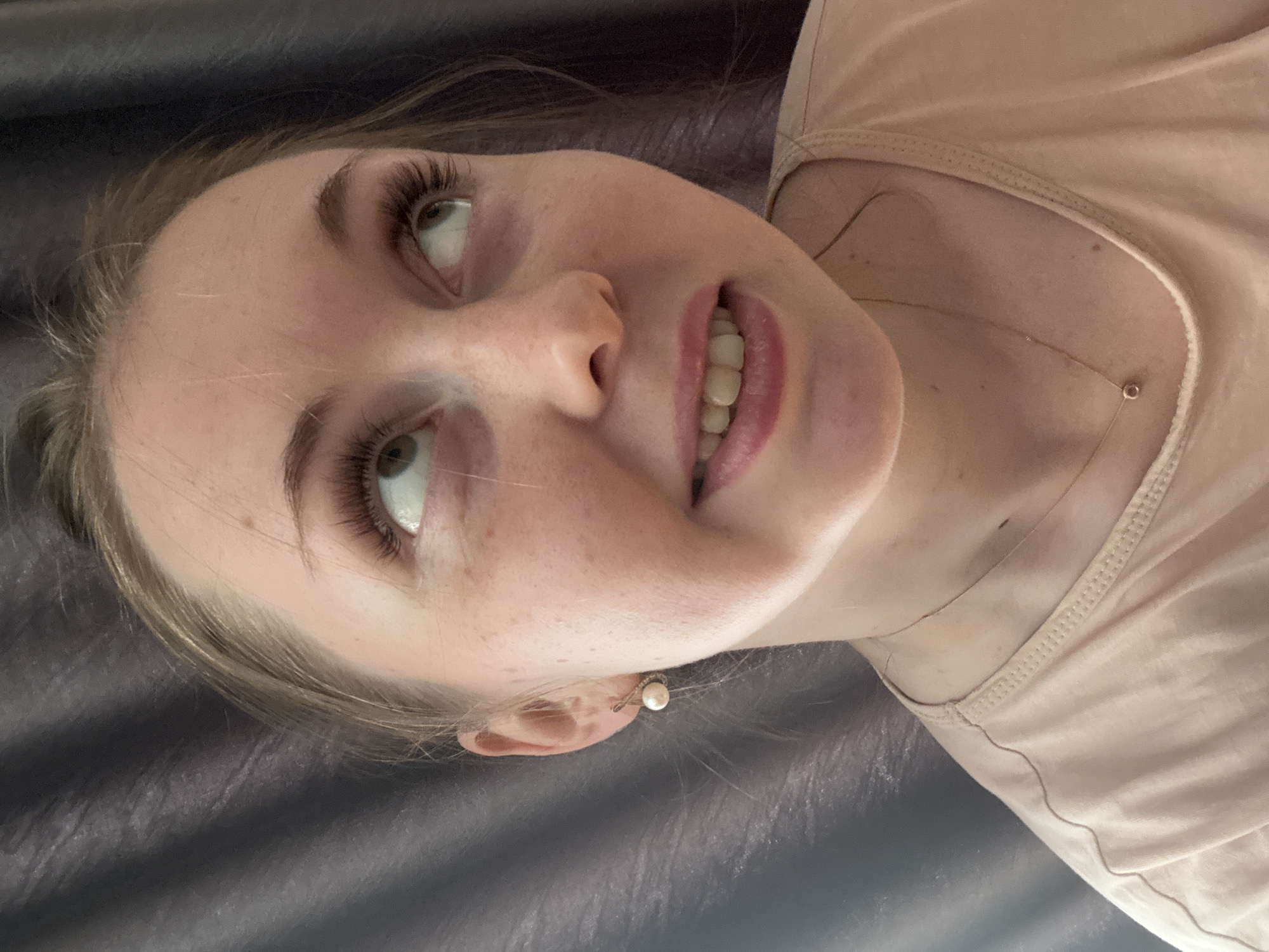 A close-up, medium-shot photograph shows a woman with fair skin, light blonde hair, and light-colored eyes. Her head is tilted slightly towards the viewer's left. She is wearing a light pink top, and a delicate gold necklace is visible around her neck. A small pearl earring is noticeable in her ear. The background appears to be a dark, textured surface, likely a bed or padded table. The setting suggests a beauty salon or personal care setting, but the exact location is not discernable.