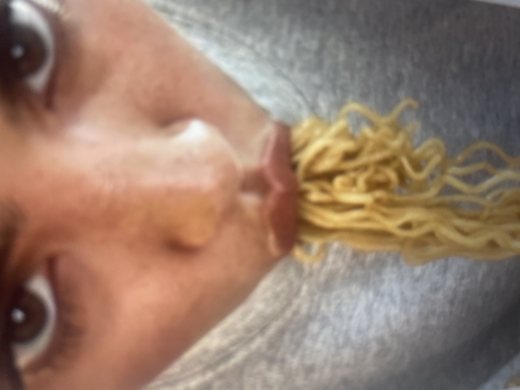 The image is a close-up, vertical shot of a person's face with noodles in their mouth. The person's eyes are visible in the foreground and background, and their mouth is open, with a cluster of curly, yellow noodles protruding from it. The skin tone of the person is light. The background is a blurred gray fabric, possibly clothing. The lighting appears to be indoor and diffused. The overall impression is one of a playful or humorous action, with the noodles creating a visually striking element against the face. There is no visible text, time of day, weather, or specific location cues. The emotions conveyed are ambiguous due to the extreme close-up and the unusual nature of the activity, but could be interpreted as playful, surprised, or a deliberate pose for a photo.