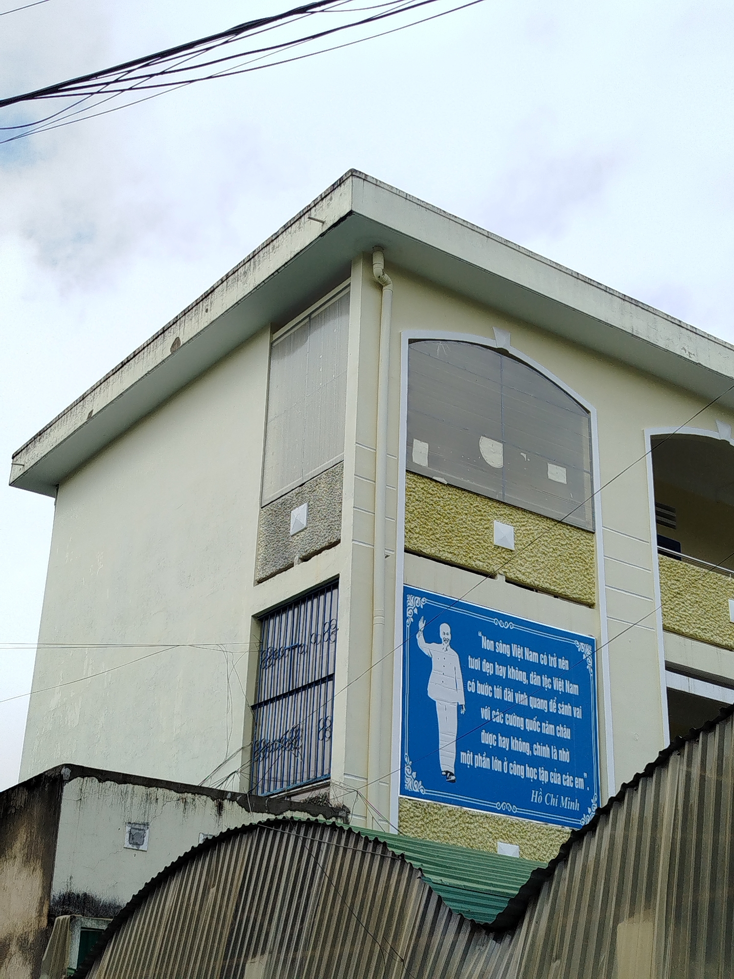 The image shows the side of a light yellow, multi-story building with a slightly weathered exterior, likely a public building such as a school or government office, located in Da Lat, Vietnam. The building has several windows, some with metal grates and others with opaque or frosted glass. A prominent blue sign with white text and a silhouette of a man in a suit is affixed to the building. The text on the sign is in Vietnamese and appears to be a quote attributed to Ho Chi Minh. The building is situated in an urban environment, with visible power lines crossing the sky in the upper portion of the frame. In the foreground, parts of other structures, including a corrugated metal roof, are visible, suggesting a dense urban setting. The sky is overcast.