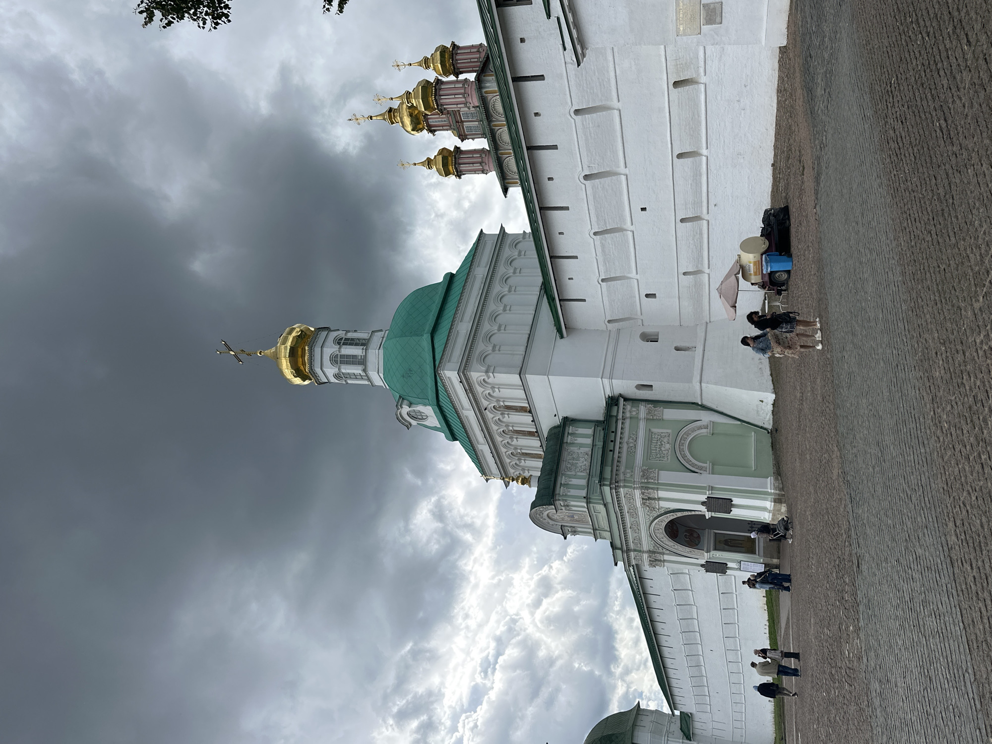 The image shows a view of the Trinity Lavra of St. Sergius in Sergiyev Posad, Russia, under a dramatic, cloudy sky. In the foreground, a cobblestone courtyard is sparsely populated with a few individuals. To the left, a group of people are walking and standing near the entrance to a white and green arched gateway, which is part of the monastery's wall. Further to the right, a small cart with an umbrella is set up, with two women standing near it. The main subject of the image is the impressive white fortress-like wall of the monastery, which extends across the frame. Several domes, some gilded and some green, are visible rising above the wall, characteristic of Russian Orthodox architecture. The overall atmosphere is somber due to the dark, overcast sky, which contrasts with the bright white of the monastery walls and the golden domes.