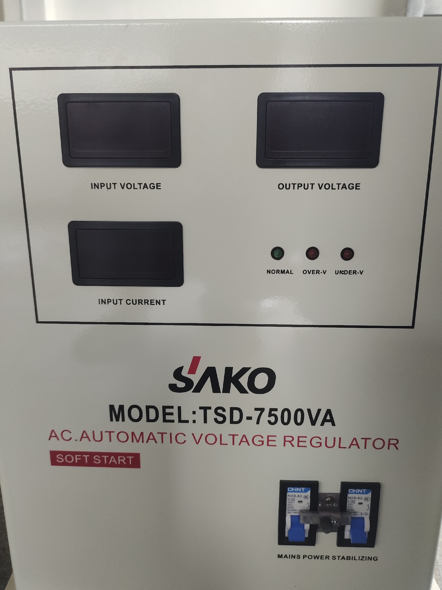 The image displays the control panel of an AC Automatic Voltage Regulator, likely indoors within a technical or industrial setting. The panel is light-colored, possibly off-white or light grey.
Prominently featured are three blank digital display screens: one labeled "INPUT VOLTAGE," another "OUTPUT VOLTAGE," and a third "INPUT CURRENT." Below the output voltage display, there are three unlit LED indicator lights: a green one labeled "NORMAL," and two red ones labeled "OVER-V" and "UNDER-V," suggesting the device is currently unpowered or in an idle state without active monitoring or fault conditions.
The brand name "SAKO" is visible in bold, stylized lettering, followed by the model number "TSD-7500VA." The device is identified as an "AC.AUTOMATIC VOLTAGE REGULATOR" and includes a rectangular red label indicating "SOFT START."
At the bottom right of the panel, two "CHNT NXB-63 C32" circuit breakers are mounted side-by-side, described by the text "MAINS POWER STABILIZING." The overall impression is of a robust piece of electrical equipment designed for power regulation and protection. No specific activity is taking place, as the image captures the static appearance of the device.
