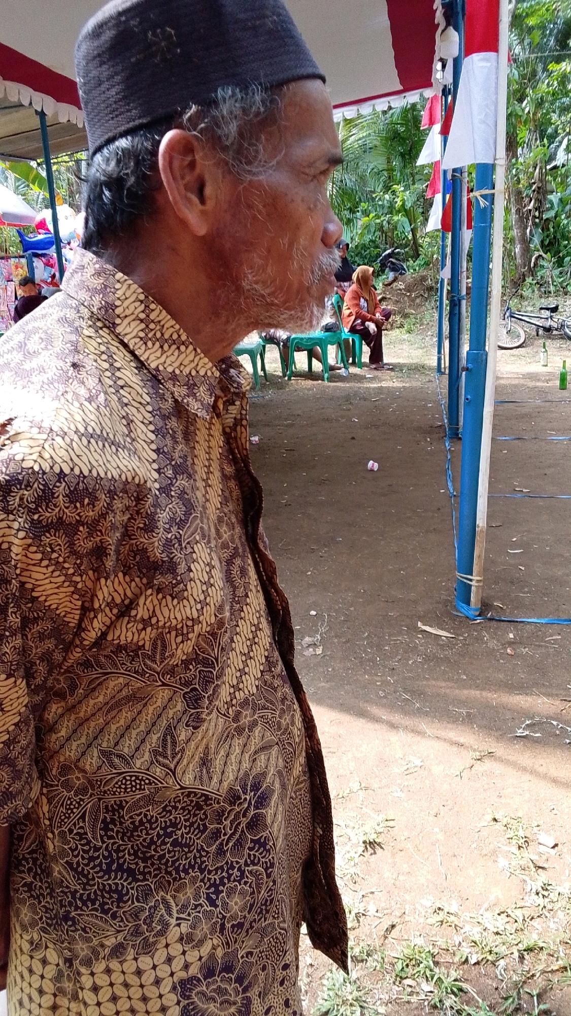 An older man with graying hair and beard, wearing a black skullcap and a patterned brown batik shirt, is shown in profile, looking to the right. He is standing outdoors, with a dirt ground and scattered debris. In the background, several people are seated on green chairs under a tent draped with red and white fabric. A bicycle is visible in the distance, and two green bottles lie on the ground. The scene appears to be a gathering or event in Parakanmanggu, Indonesia.