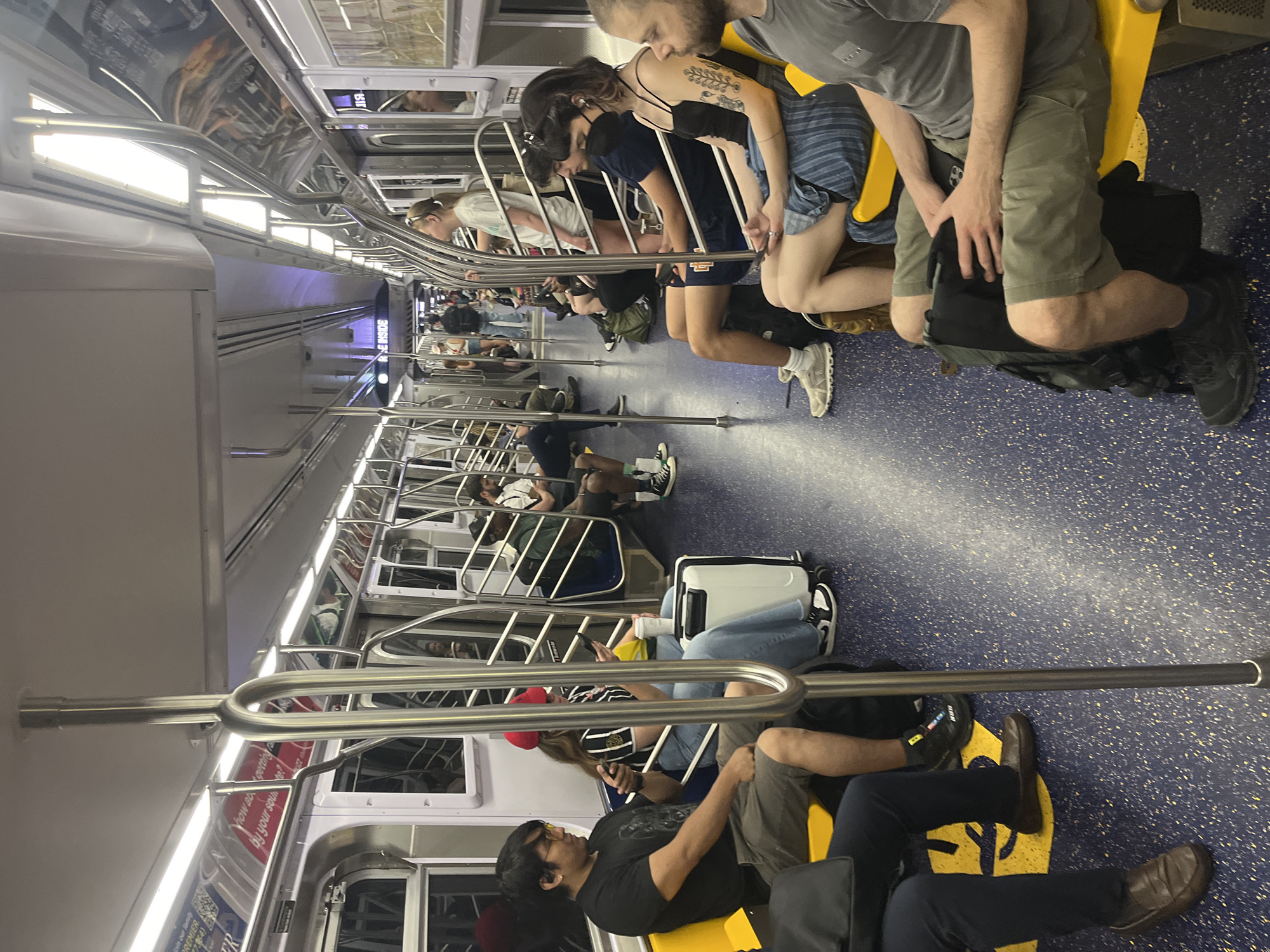 The image captures the interior of a subway car, likely in New York City, as indicated by the location context. The scene is populated with several people, some seated on yellow subway seats and others standing. The time of day is indeterminate, and there are no clear weather cues.
The subjects are diverse, with individuals of various ages and ethnicities. They appear to be commuters, with some engaged in personal activities like using their phones, while others simply rest. There is a sense of normalcy and routine associated with public transit.
Visible objects include luggage, such as a white rolling suitcase, as well as personal bags and backpacks. The subway car itself is characterized by its metal railings, seating, and illuminated interior. The floor is a speckled blue, and advertisements are visible on the walls, though much of the text is partially obscured or illegible in the main image.
Some of the visible text includes "PREMIER" and a partial phrase that seems to be "Yhow ab getting by your soute?". Another sign reads "REINSIDE". These appear to be advertisements or public service announcements within the subway car.
The overall impression is that of a typical scene inside a busy public transportation vehicle, where people are in transit, each absorbed in their own journey.