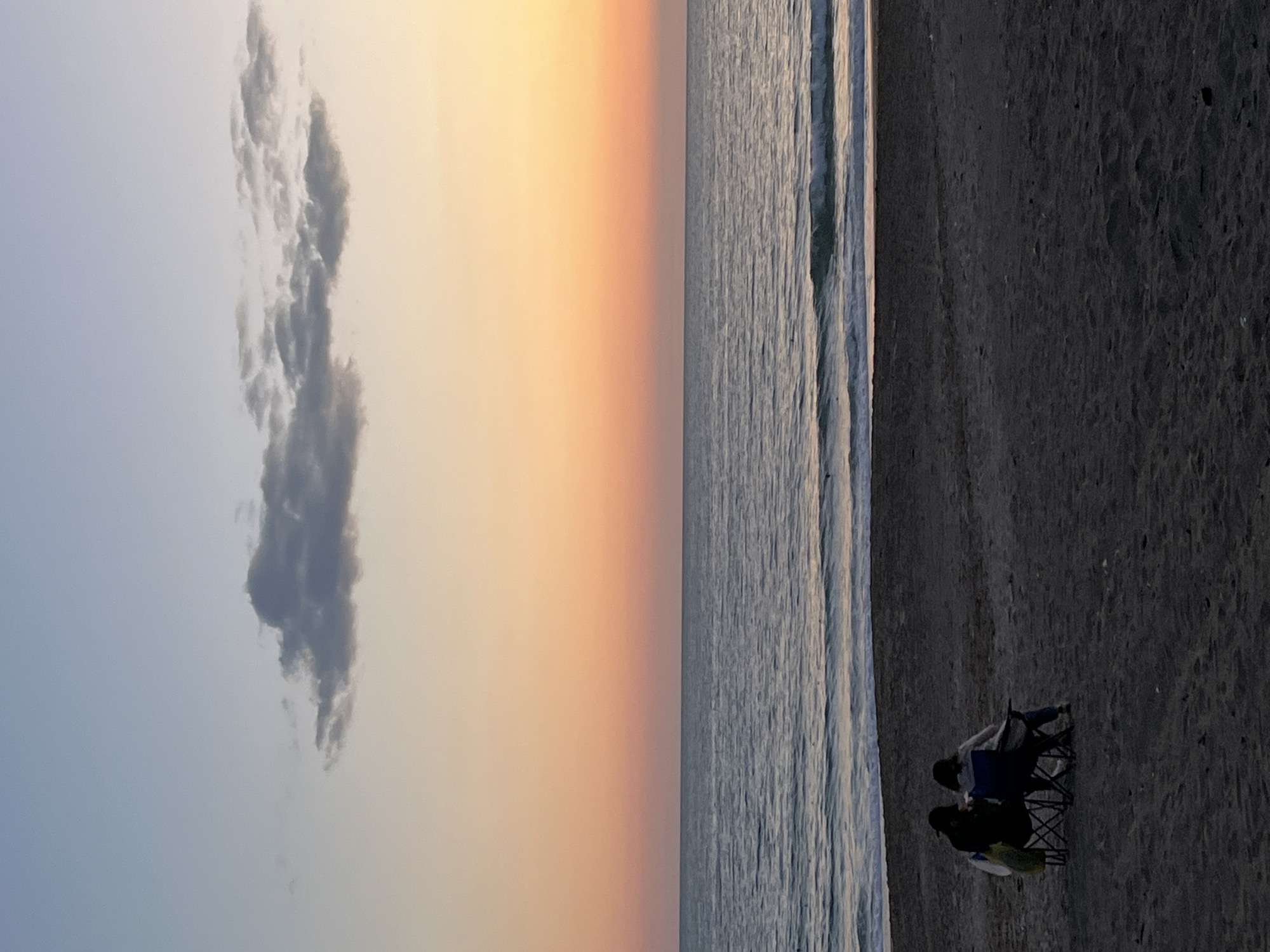 Two people sit in folding chairs on a beach, with the ocean to their left. The sky shows a gradient from light blue to orange. A large cloud hangs in the sky. The scene is at a beach in Alanya, Turkey.