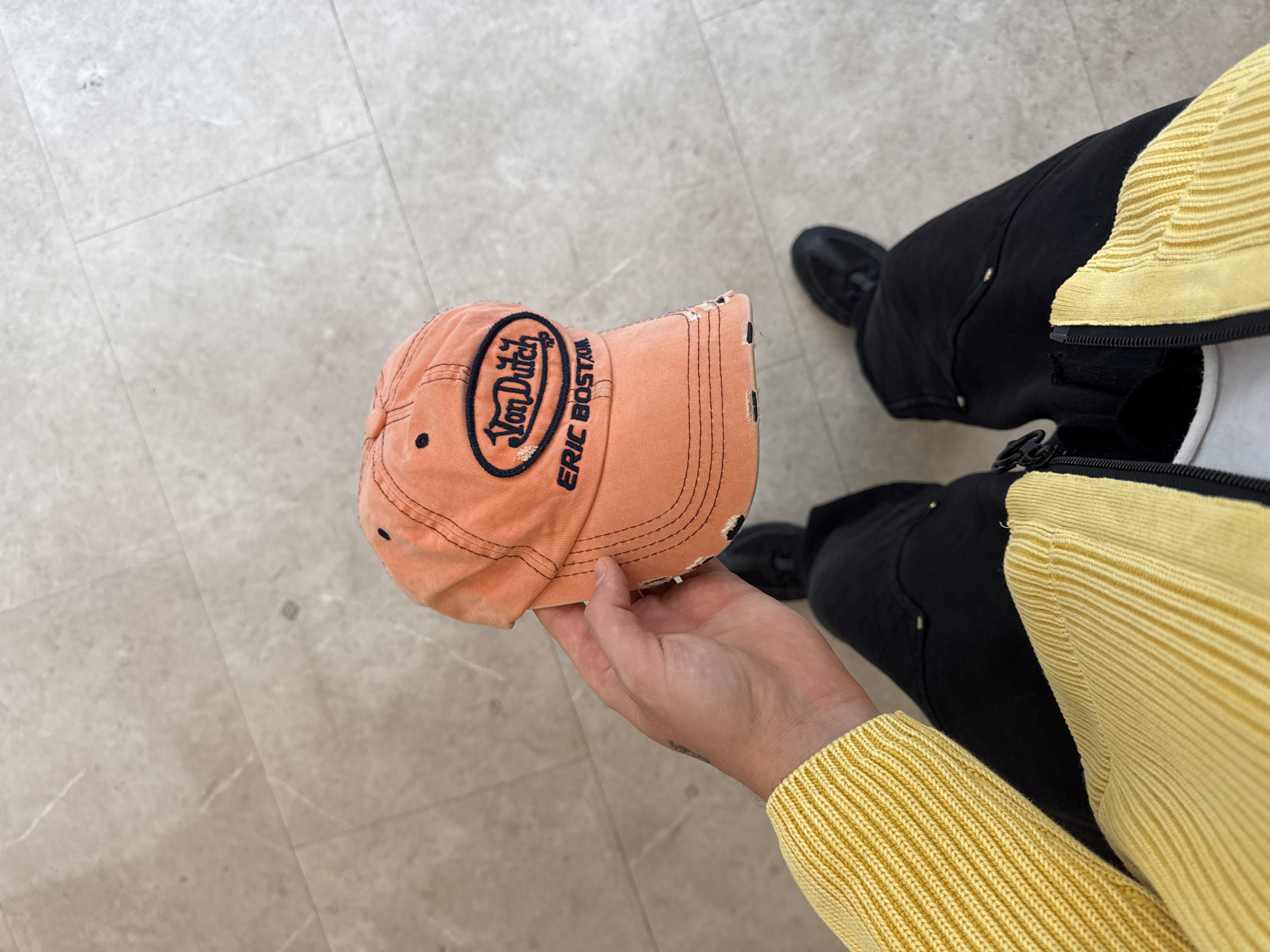 A top-down shot shows a person holding an orange baseball cap with a black "Von Dutch ERIC BOSTON" logo. The person is wearing black pants and a yellow sweater. Part of the black shoe is also visible. The background appears to be a beige-tiled floor. There is no indication that the photo was taken in London, United Kingdom.