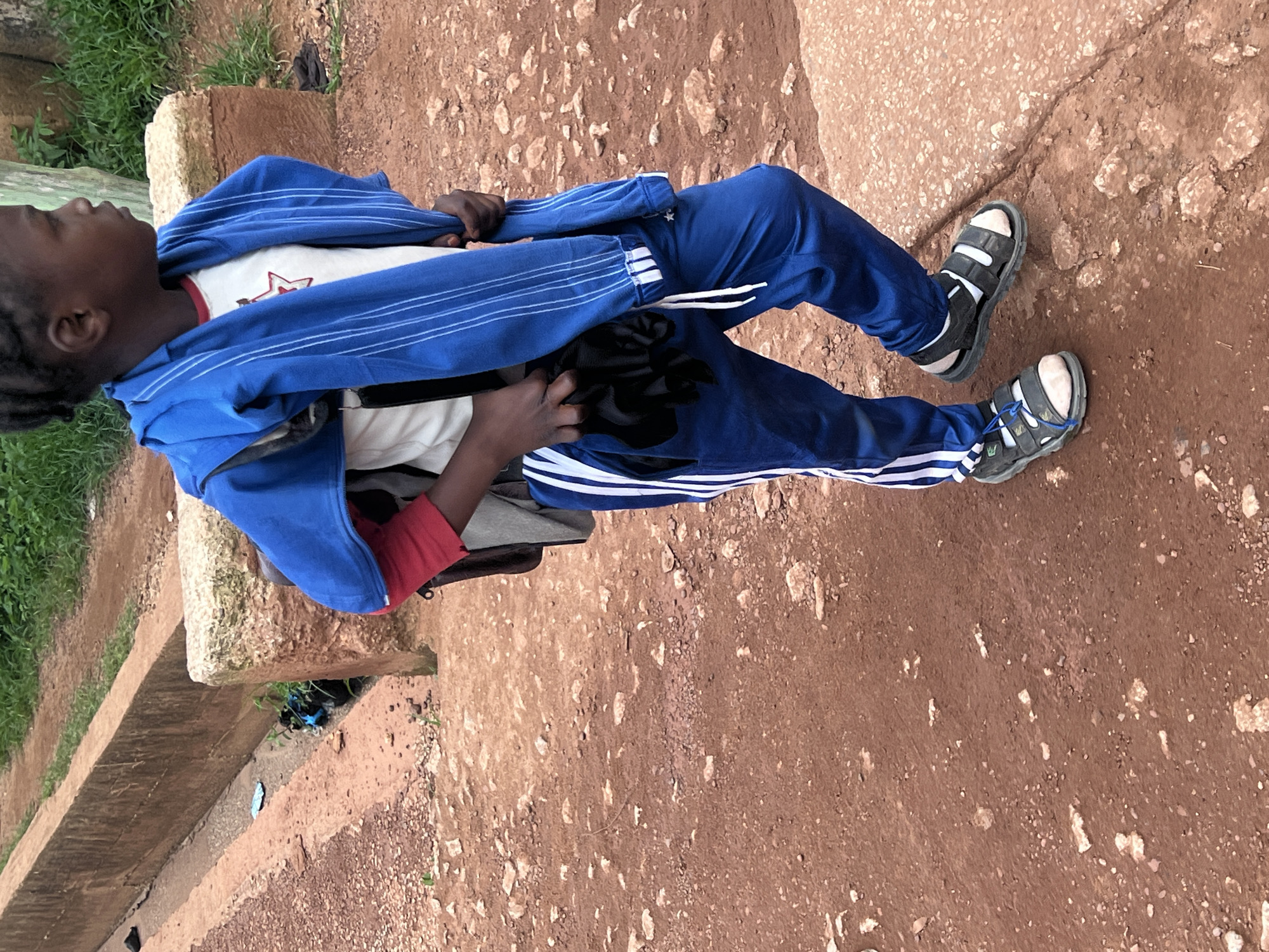 The image depicts a young person standing on a dirt path in Durumi, Nigeria. The individual is wearing a blue hooded jacket draped over their shoulders, a white t-shirt with a red star graphic, blue track pants with white stripes, and black and white sandals. They are carrying a backpack and holding a black object, possibly gloves, in their left hand. In the background, a concrete drainage channel is visible, with some greenery and debris along its edge. The ground is composed of reddish-brown dirt and small stones. The overall scene suggests an outdoor, possibly urban or semi-urban, environment.