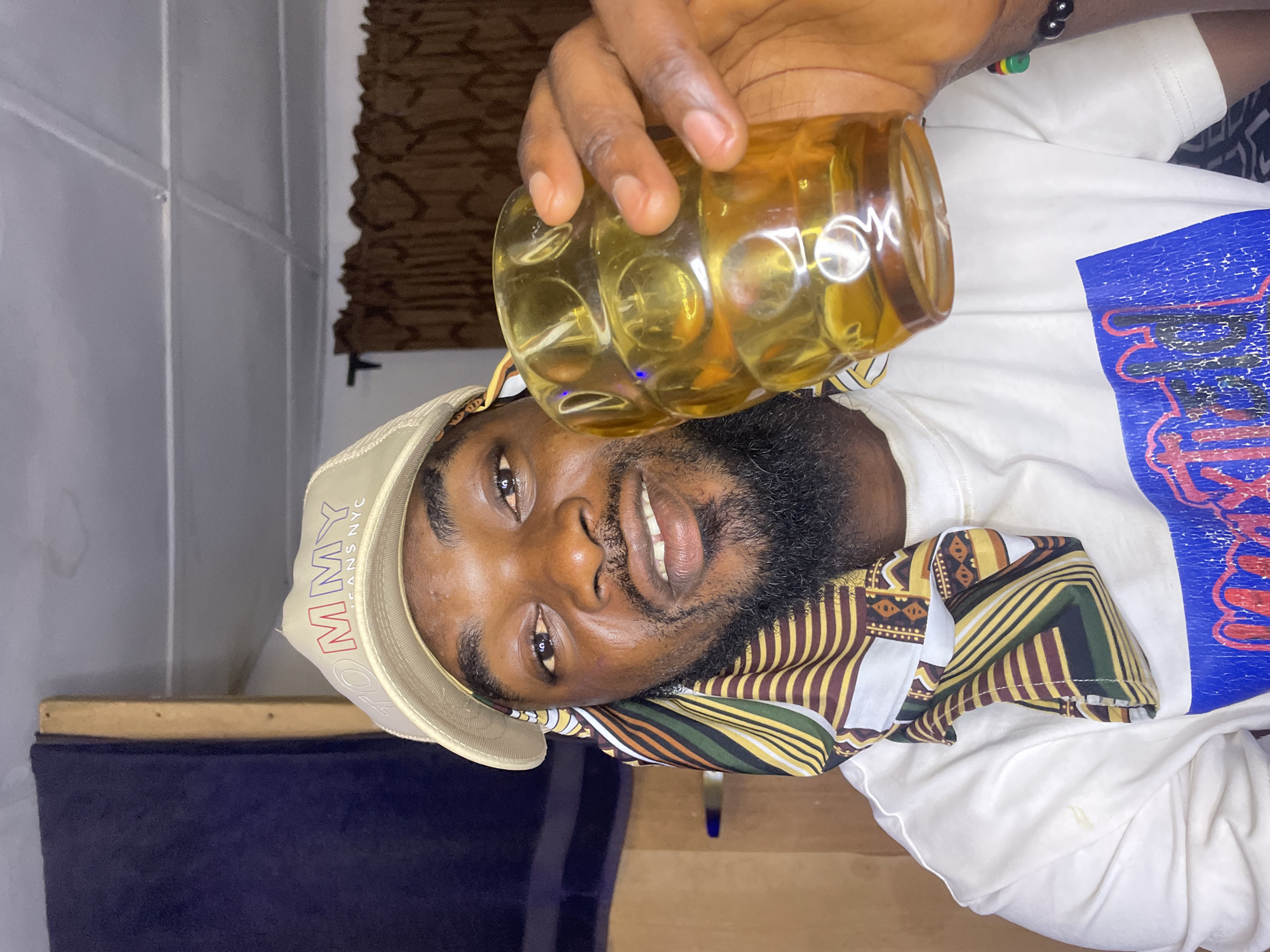 A medium shot depicts a man with dark skin, a short beard, and a bald head. He is wearing a light-colored Tommy Jeans hat and a patterned scarf, and a white t-shirt with a graphic on the sleeve. He holds a yellow plastic jar with liquid, perhaps juice. He is smiling and looking at the camera. The location is unknown, but the prompt suggests Jos, Nigeria. The background includes a dark-blue couch and a gray wall with a hanging brown-and-black patterned textile.
