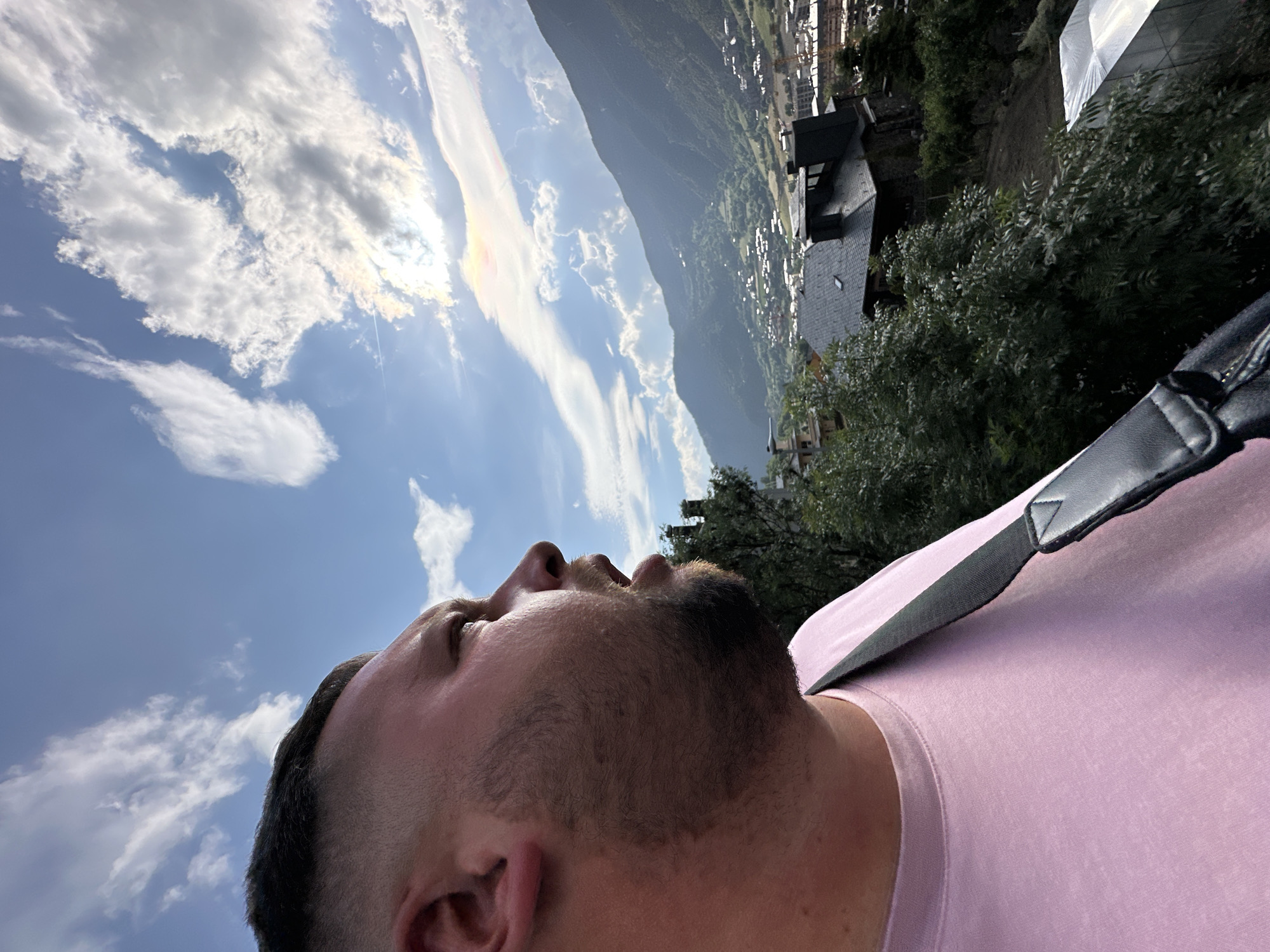 The image, originally rotated clockwise, features a close-up, low-angle profile shot of a man from the chest upwards, looking intently towards the sky. He has short, dark hair and a neatly trimmed beard, and is wearing a light pink t-shirt. A black strap, possibly from a bag or camera, is visible diagonally across his right shoulder.
Above and behind the man, a vibrant blue sky is filled with large, white cumulus clouds. The sun is prominently visible through a thinner cloud layer, creating a bright glare and casting a subtle iridescence with rainbow hues on the edges of some clouds. A faint contrail from an aircraft is also discernible in the sky.
The background reveals a sprawling mountainous landscape. Below the expansive sky and clouds, parts of a town or village are visible in the valley, characterized by buildings with dark roofs nestled among abundant green trees and foliage. The distant mountains are imposing, blanketed with verdant greenery.
The man appears to be observing the sky and the scenic view of the valley and mountains. The time of day is clearly daytime, characterized by bright sunlight. The overall setting points to a high-altitude natural environment. The location context for this scene is Encamp, Andorra. No visible text is present in the image.