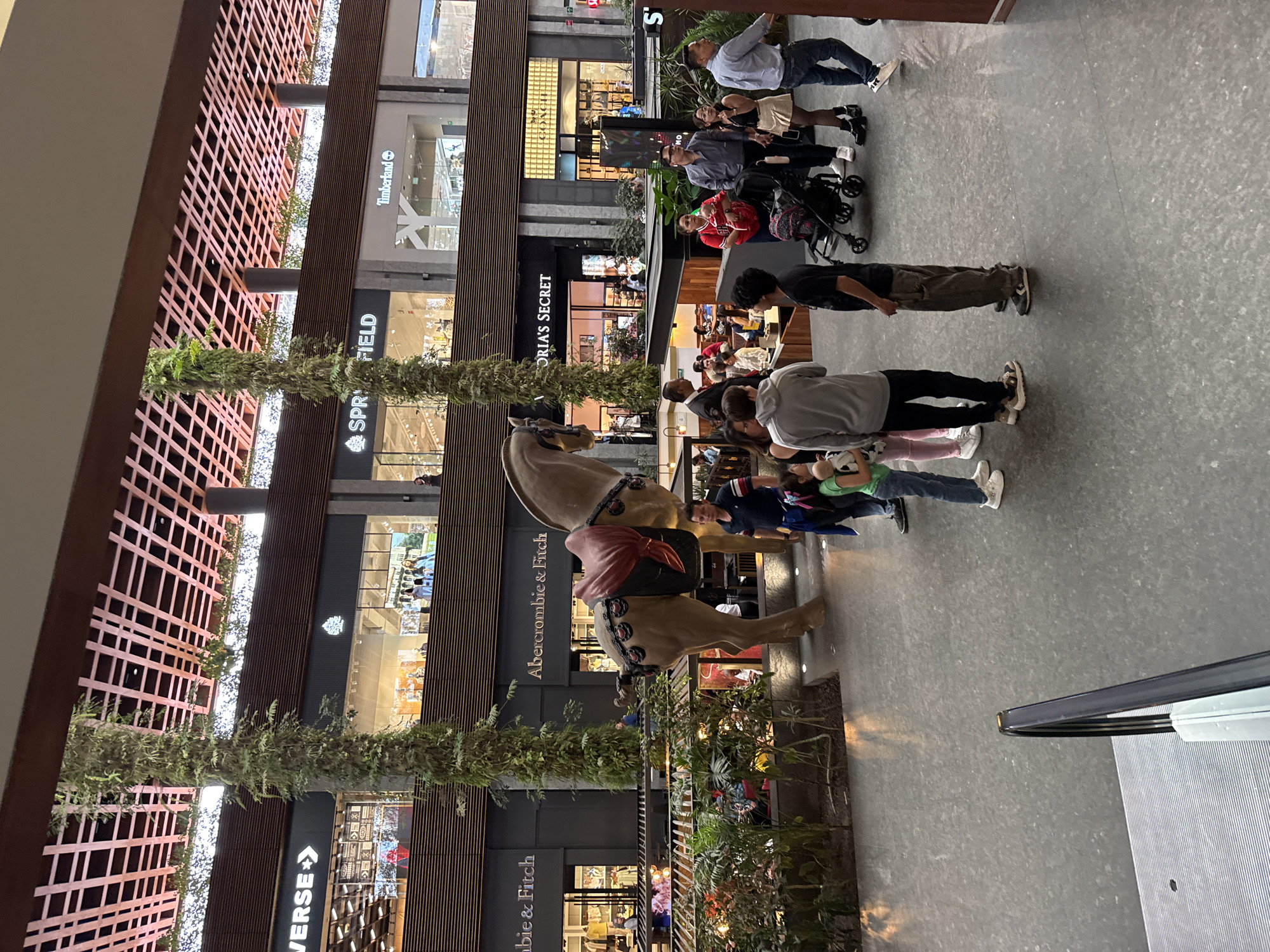 This photograph captures a multi-level indoor mall environment. Several stores are visible, including Abercrombie & Fitch, Converse, Springfield, Timberland, and Victoria’s Secret. Numerous individuals are present, engaged in activities such as shopping, dining, and walking. The environment includes decorative greenery and a large horse sculpture that is surrounded by visitors. The overall atmosphere suggests a busy commercial setting. The location is presumed to be in the city of Puebla, Mexico.