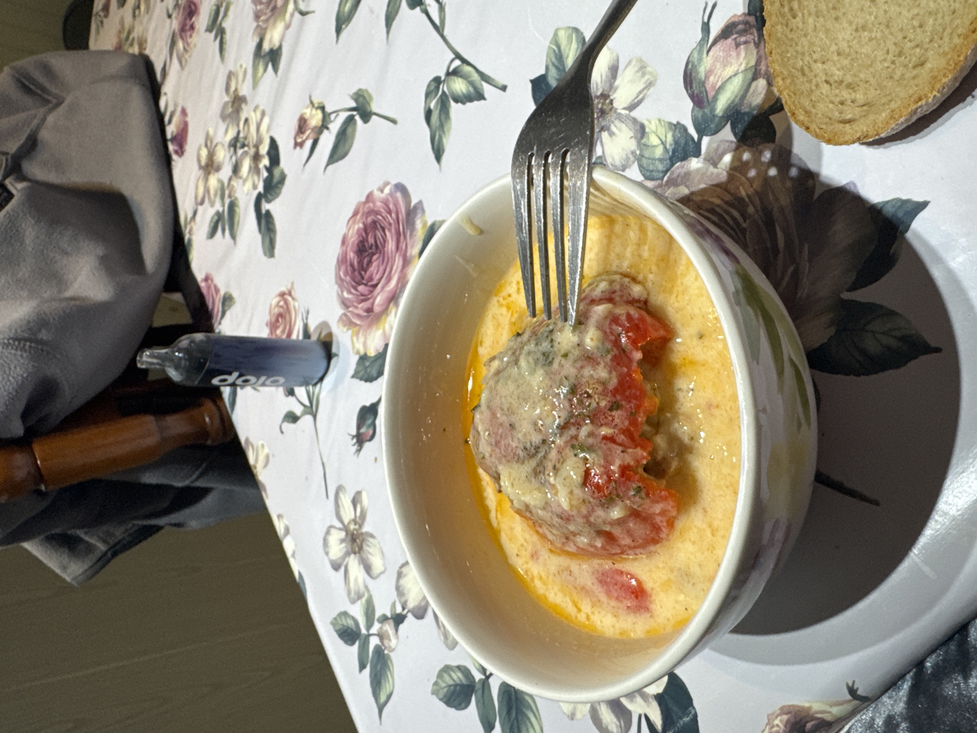 A close-up, eye-level shot captures a bowl of food on a table covered with a floral tablecloth. The bowl contains a dish that appears to be a stuffed pepper, covered in a creamy sauce with visible chunks of red pepper and herbs. A fork is inserted into the food, suggesting it is being eaten. To the right of the bowl, a slice of bread rests on the tablecloth. In the background, a blue vape pen lies on the table, next to a dark grey cloth item draped over a wooden object. The overall scene suggests a meal being consumed in a domestic setting. The location is identified as the city of Armagh, United Kingdom.