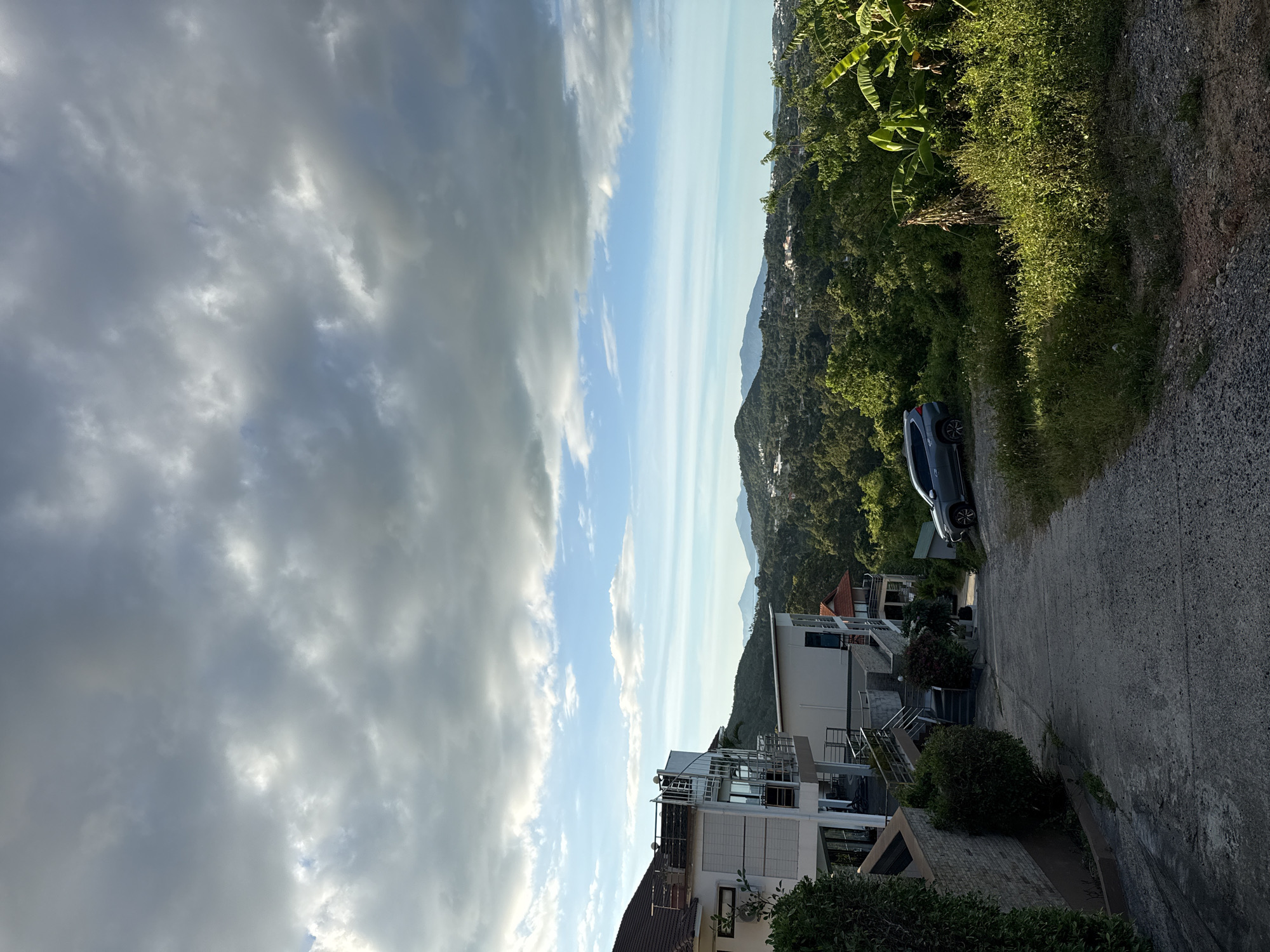 This is a photograph of a hilly, tropical landscape with modern architecture. The scene appears to be a residential area, possibly a resort or a collection of villas, situated on a hillside overlooking a distant body of water and mountains. In the foreground, a paved road curves uphill, with a dark-colored car parked on the right side. Several multi-story buildings with white exteriors and balconies are visible, some featuring contemporary designs with glass railings. Lush green vegetation, including banana trees and dense foliage, covers the slopes, creating a vibrant, natural backdrop. The sky is a mix of blue with scattered, dramatic clouds that suggest either approaching or receding weather. The lighting indicates it is late afternoon or early evening, with the sun casting long shadows and highlighting the textures of the clouds and landscape. The image conveys a sense of serene, upscale living in a tropical setting.