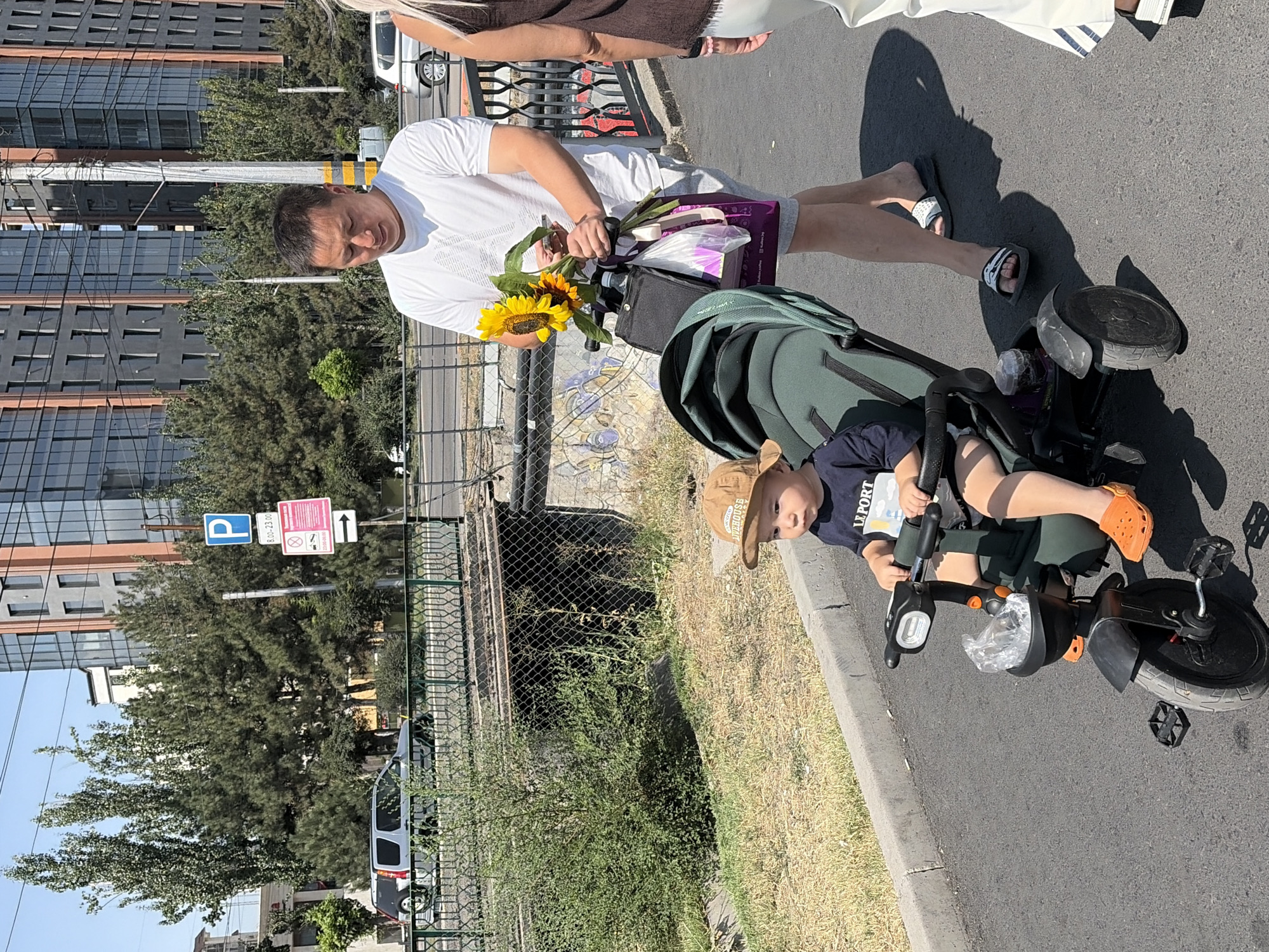 The image shows a man holding a bouquet of sunflowers and another person, possibly an adult, standing behind him. A young child is seated in a stroller or trike next to them. The scene appears to be outdoors, on a paved surface that looks like a sidewalk or street, with a grassy area and trees in the background. There are also buildings visible, suggesting an urban or suburban environment.
The man is wearing a white t-shirt and shorts, and the child is wearing a brown hat and a dark shirt. The child's legs are visible, and they are wearing orange shoes. The man is holding the flowers with both hands, and his head is tilted down as if looking at them or talking to the child. The overall atmosphere seems casual and summery, with bright sunlight casting shadows.
Visible text includes "LE PORT" on the child's shirt and signs on a pole in the background, one of which appears to be a parking sign with time restrictions. The other sign is pink and has some text and symbols that are difficult to read. The weather appears to be sunny and warm.