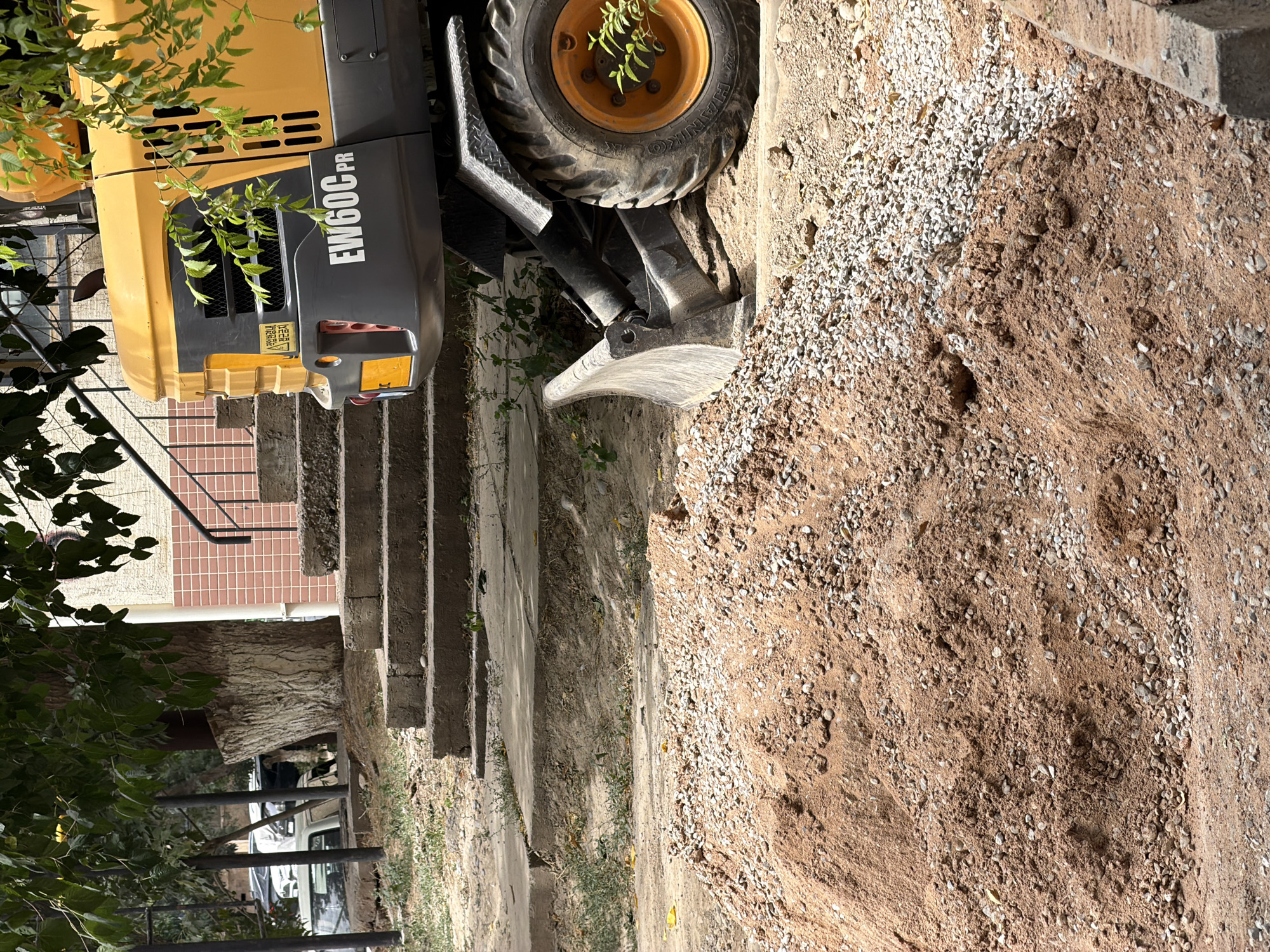 A yellow excavator with the model number EW60C PR is parked on a paved surface near a pile of sand and gravel. Behind the excavator are concrete steps leading up to a building with a pink tiled facade and black metal railings. Trees with green leaves are visible in the background, casting shadows on the scene. The environment appears to be an urban outdoor setting. There are no people visible in the image. The scene is located in Shymkent, Kazakhstan.