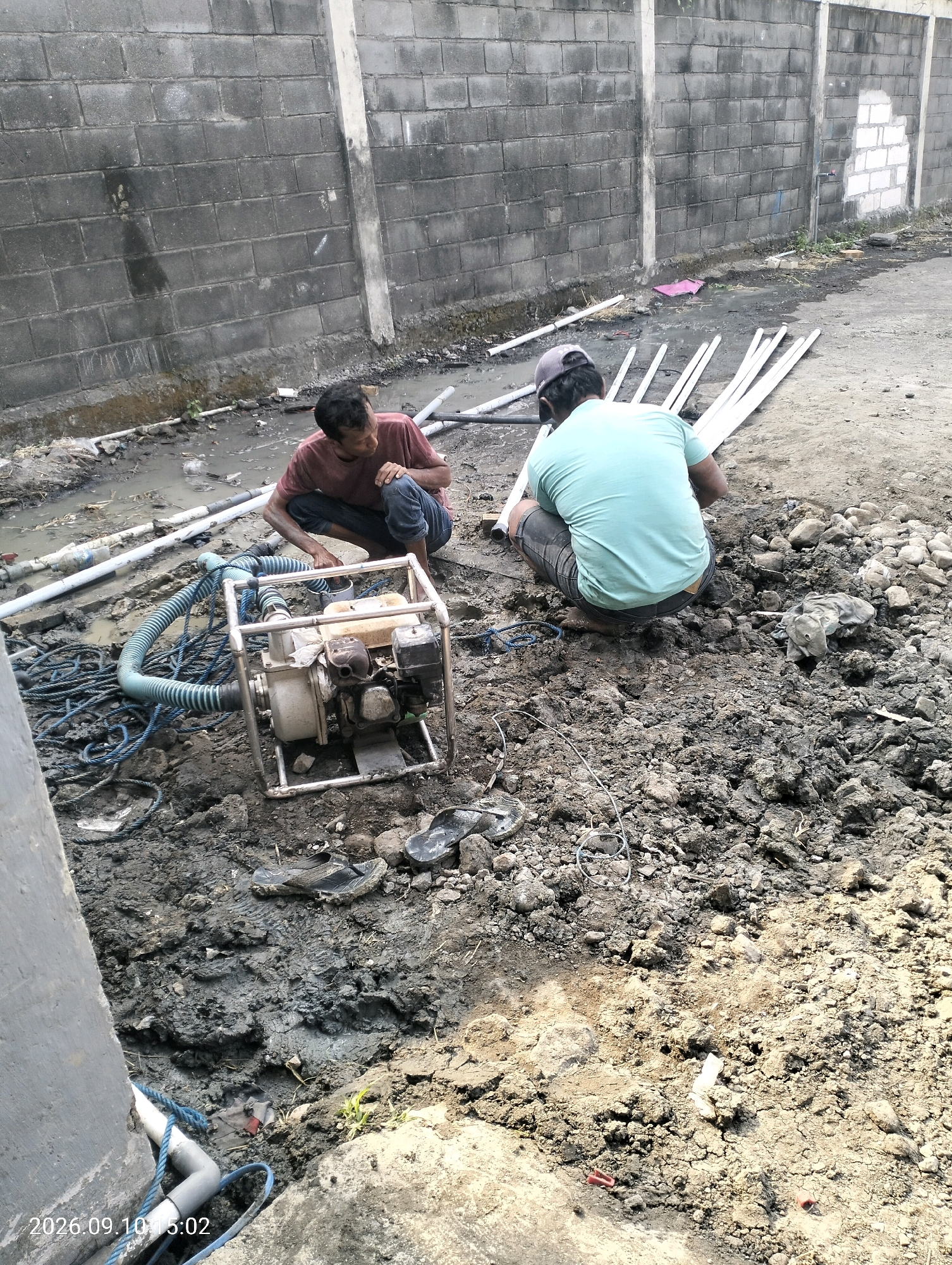 Two men are working in a muddy construction area in Bambe, Indonesia. One man, wearing a maroon shirt and dark pants, is crouching near a water pump and a coiled blue rope. The other man, wearing a light blue t-shirt and dark shorts, is kneeling further away, facing the wall. The ground is uneven and covered in mud, rocks, and construction debris. Several white pipes are laid out on the ground, and a hose is connected to the water pump. A grey cinder block wall forms the background, with some vegetation growing near its base. The scene appears to be an outdoor construction site where drainage or water management is in progress.