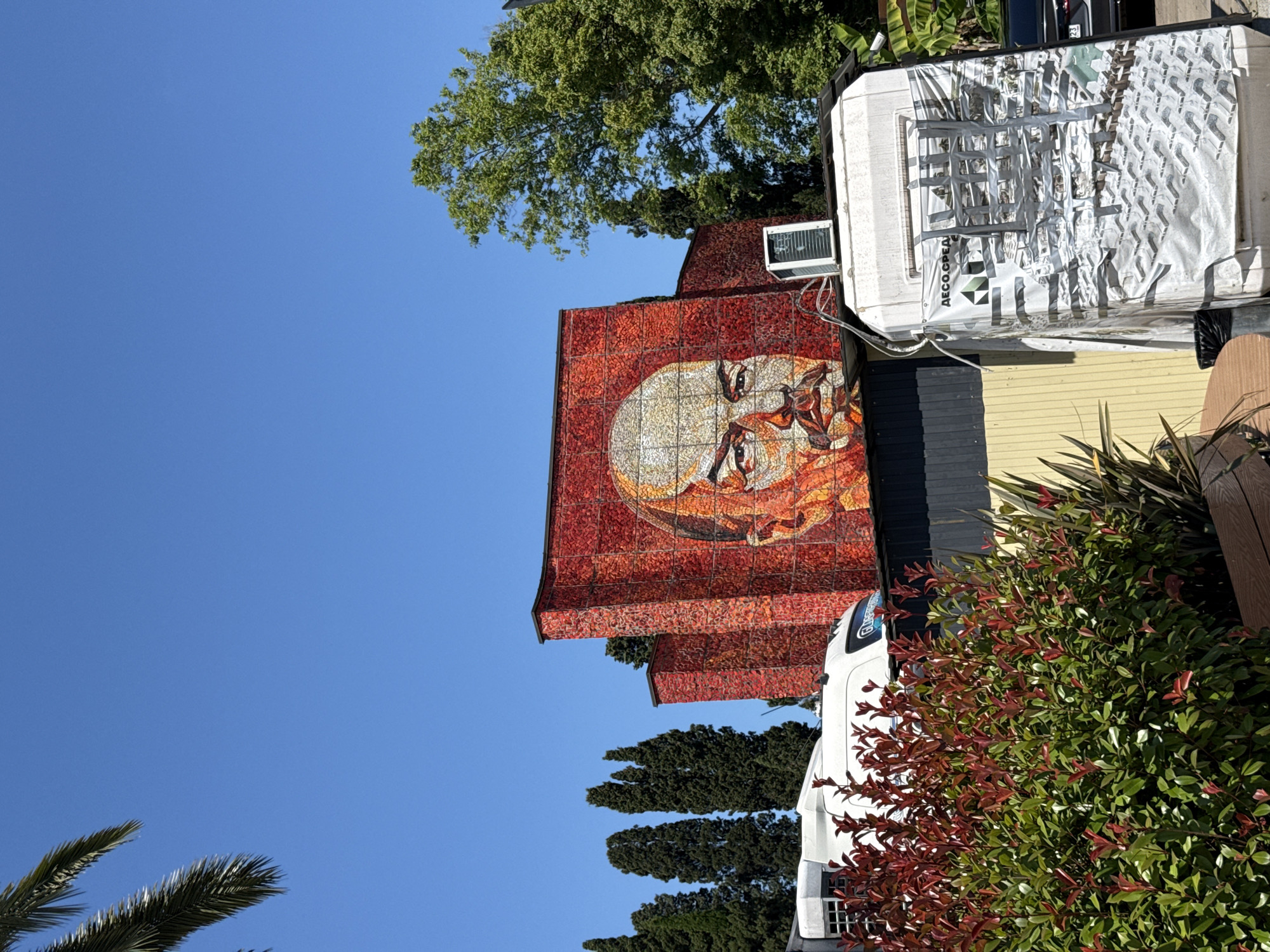 The image depicts a large mosaic of Vladimir Lenin's face on the side of a building in Sochi, Russia. The mosaic is predominantly red and orange, with Lenin's face rendered in shades of yellow and white. The building itself appears to be curved and has a textured, reddish exterior, likely made of small tiles or bricks.
In the foreground, there is lush green foliage with some red leaves, partially obscuring a white van. The van has a large white tarp attached to its side, which is covered in tape and appears to have some text and images on it, including the Cyrillic letters "ДЕСО.СРЕД". Above the van, on the roof of a smaller structure, an air conditioning unit is visible.
Behind the mosaic building, several tall, dark green cypress trees stand against a clear blue sky. To the right of the mosaic, a large, leafy green tree is visible. In the lower right corner, a part of a dark-colored car can be seen. The overall scene suggests an outdoor setting on a sunny day. The mosaic of Lenin is a prominent cultural or historical landmark in Sochi.