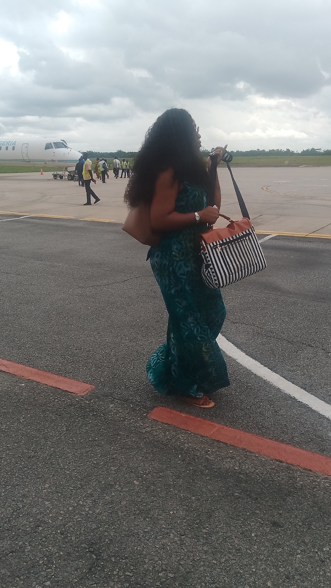 The image shows a woman walking on an airport tarmac under a cloudy sky. She is wearing a long, teal, patterned dress and carrying two bags: a tan tote bag over her left shoulder and a striped crossbody bag with brown accents in her right hand. She has dark, curly hair and is wearing sandals. Several other people are visible in the background near a white airplane with "IGERIA" written on its side, suggesting the location is Nigeria. Some of these individuals appear to be airport personnel wearing high-visibility vests. The scene appears to be in the daytime, with overcast weather indicated by the cloudy sky. The tarmac is marked with white and red lines.