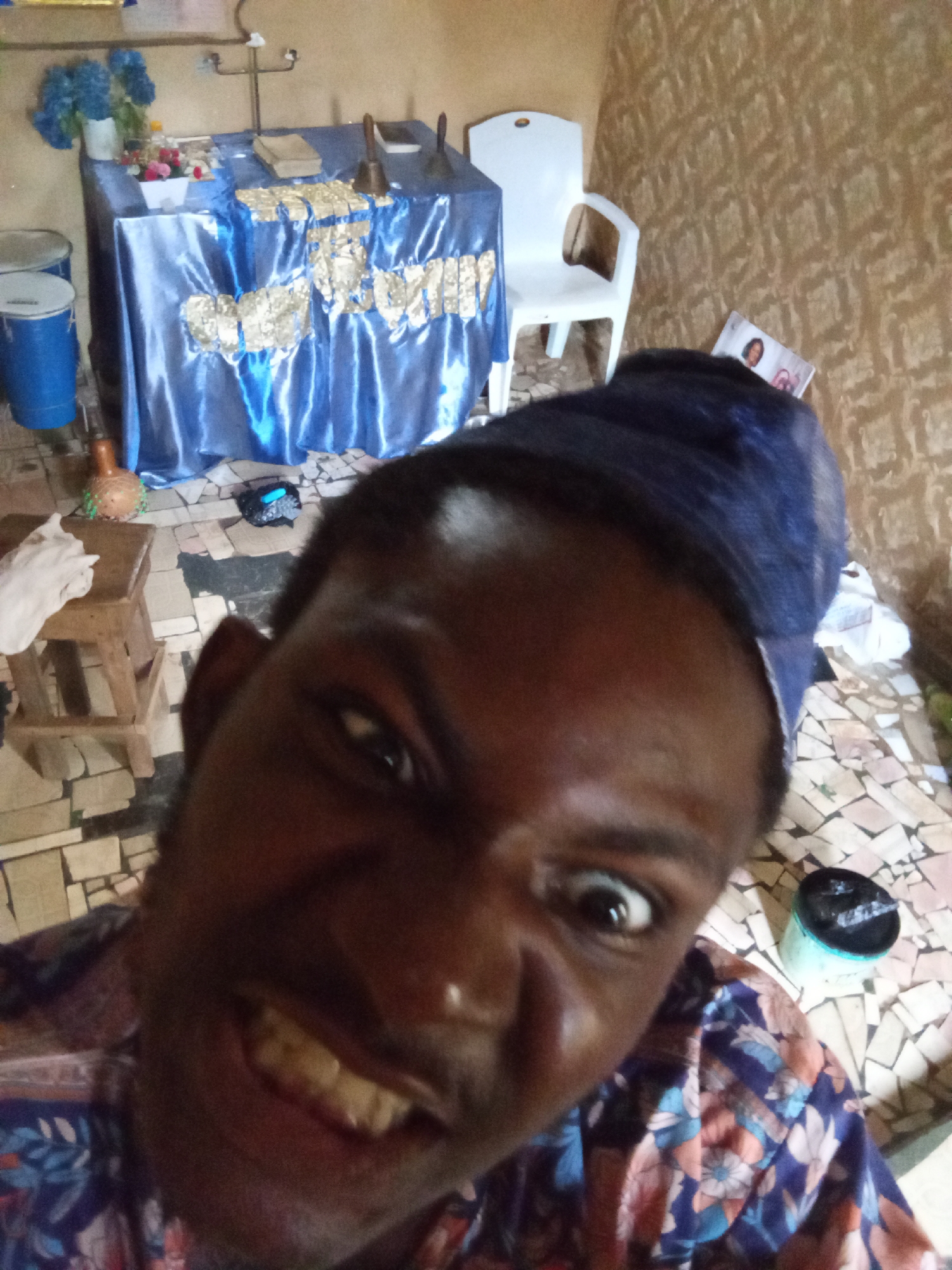 A close-up selfie captures a dark-skinned man with a blue head covering. He's pulling a comical face, baring his teeth and widening his eyes. The background reveals a room that appears to be a place of worship, with a table draped in blue fabric adorned with gold-colored decorations, and a cross-shaped metal object. A white chair stands nearby. A blue bucket sits on the patterned tile floor. The location is unknown.