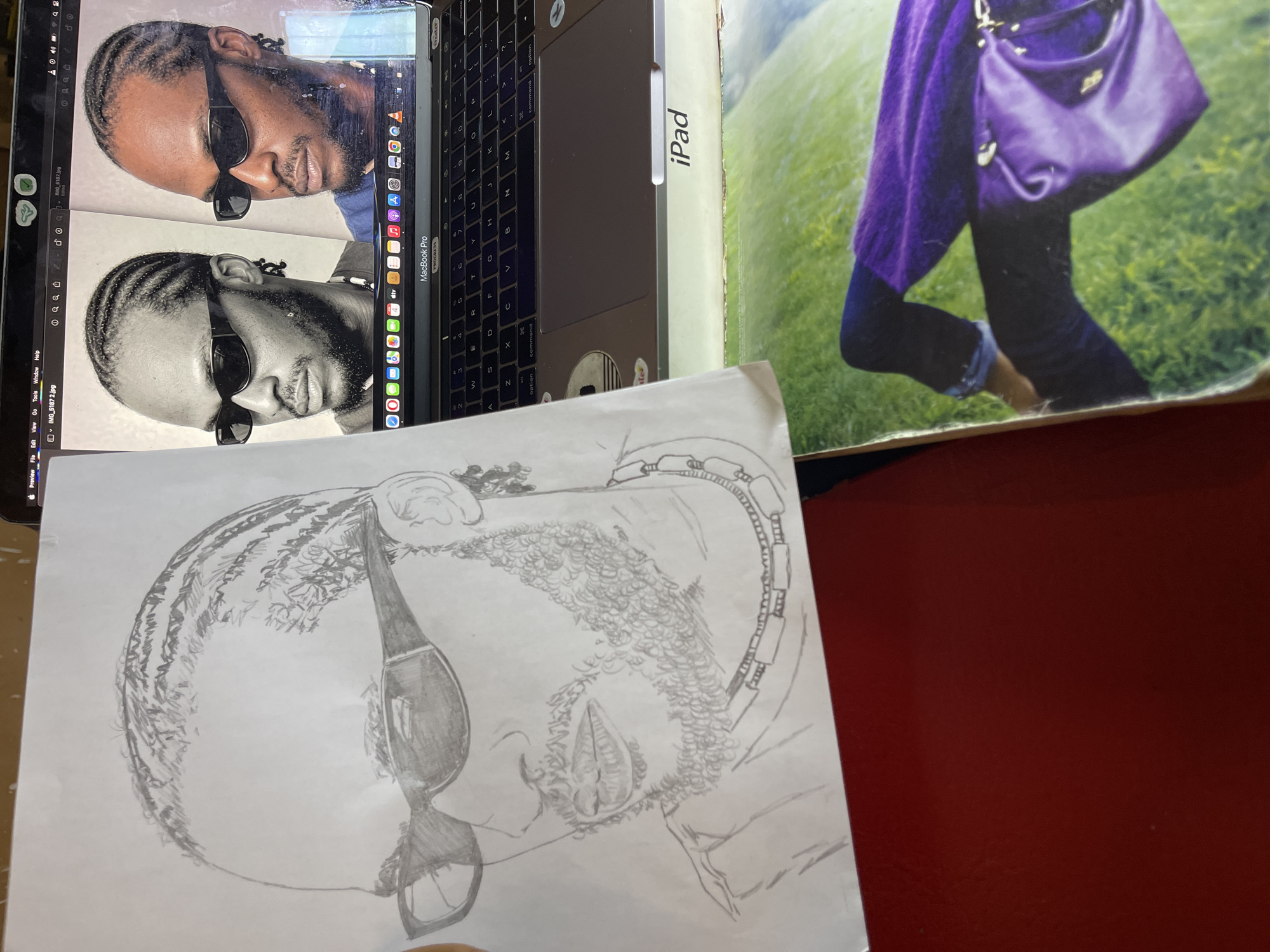 The media file captures an indoor scene, likely a workspace, with a focus on an artistic activity.
In the center, a silver MacBook Pro laptop displays two images of a man in portrait orientation. The upper image is in color, showing a man with dark braided hair, a beard, and black sunglasses, looking slightly upward and to his right. The lower image is a grayscale version of the same man, also wearing sunglasses and braids, with a similar expression. Both images are open in the "Preview" application, with menu items "File Edit View Go Tools Window Help" visible, along with filenames "IMG_5187 2.jpg" and "IMG_5187 1.jpg". The laptop itself shows "MacBook Pro" and "PROGRESS" printed on its surface.
Below the laptop screen, a pencil drawing on white paper depicts a man's head, similar to the man on the screen, shown in profile. The drawing features braided hair, sunglasses, and a textured beard, rendered with pencil strokes. The drawing is positioned as if being worked on, likely using the digital images as reference.
To the right of the laptop, a partial view of an "iPad" box or device is visible. Further to the right, a large cropped image (possibly a magazine or poster) shows the lower body of a person from the waist down, wearing a textured purple sweater or shawl, dark pants, and carrying a purple shoulder bag with gold hardware. The background of this image is vibrant green grass. The surface supporting these items is a dark reddish-brown table.
The primary activity depicted is the creation of a pencil drawing, with the digital images on the laptop serving as the subject reference. The scene is illuminated by indoor lighting. The general context for this location is Jos, Nigeria.
