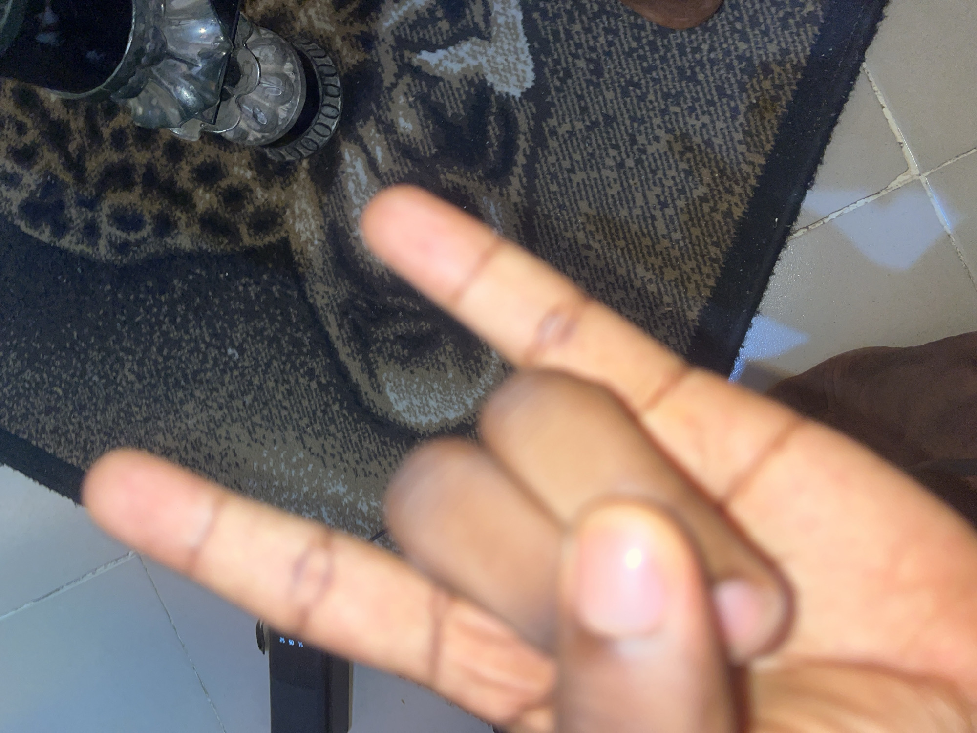 A close-up shot shows a person's left hand making a "rock on" gesture, with the index and pinky fingers extended upwards and the middle and ring fingers curled down. The hand is positioned over a patterned rug with a leopard print design and a black border. A light-colored tiled floor is visible in the lower portion of the image. The background is out of focus, but it appears to be an indoor setting. No specific location in Jalingo, Nigeria can be identified from the image.