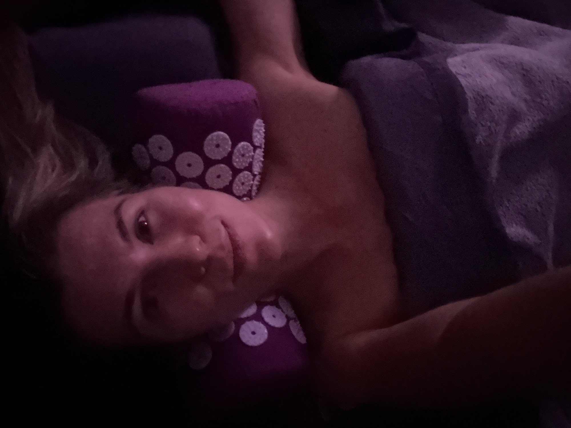 A woman lies on a dark surface, partially covered by a textured, gray-purple blanket. Her head rests on a purple pillow adorned with white, circular designs. The woman's face is visible, and she appears to be looking directly at the viewer. The location is unspecified, and there is no indication of a city or specific environment.