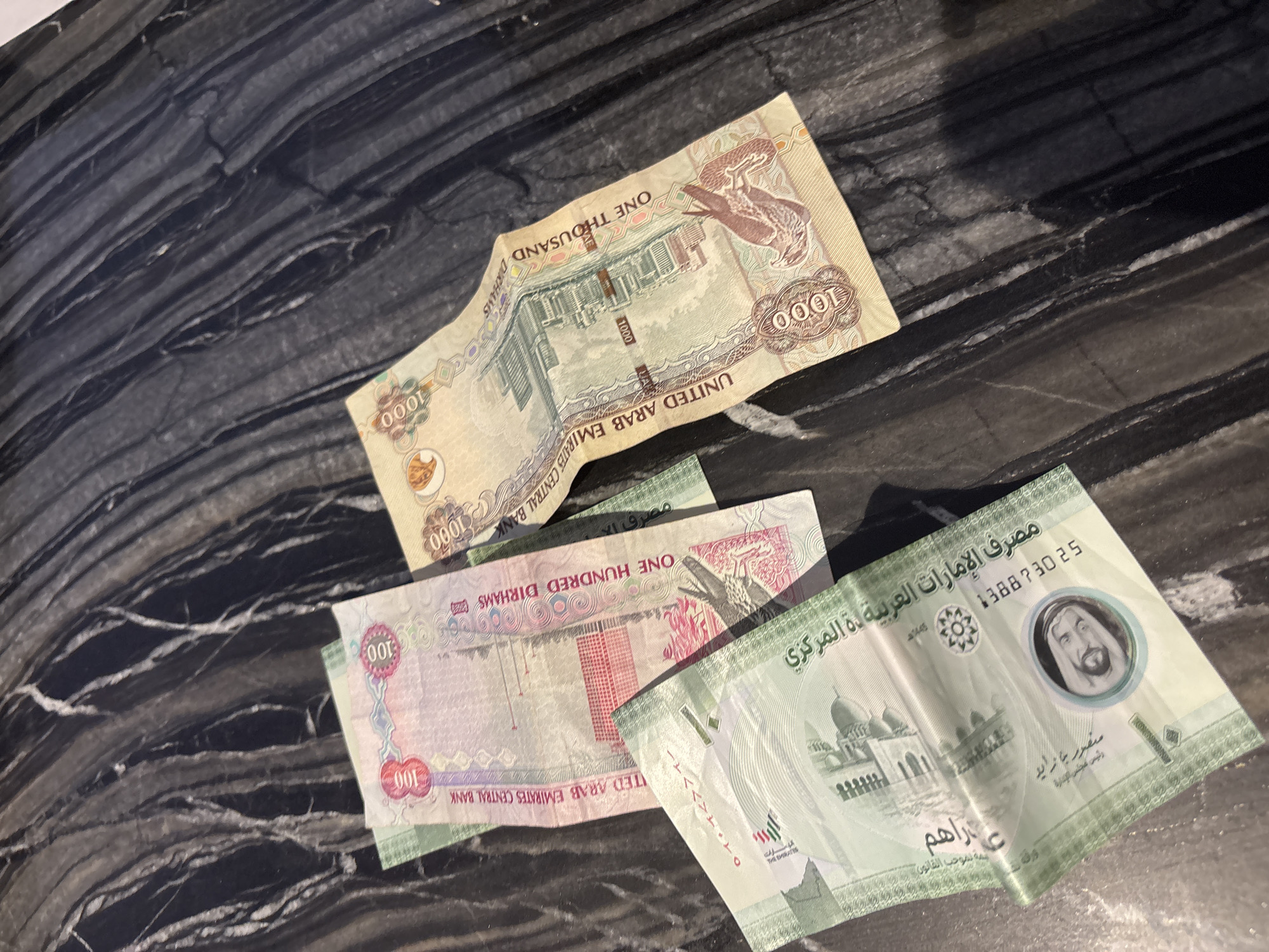 The image displays three United Arab Emirates (UAE) dirham banknotes scattered on a dark, glossy, marble-patterned surface. One banknote, predominantly pink and white, shows the denomination "100" and text reading "UNITED ARAB EMIRATES CENTRAL BANK" and "ONE HUNDRED DIRHAMS". Another banknote, yellow and beige, is folded and shows the denomination "1000" and text reading "UNITED ARAB EMIRATES CENTRAL BANK" and "ONE THOUSAND DIRHAMS". A third banknote, mostly green, is partially visible and shows text in Arabic and the denomination "100". The lighting suggests an indoor environment, likely a table or countertop. No specific landmarks or indications of Dubai's city are present.