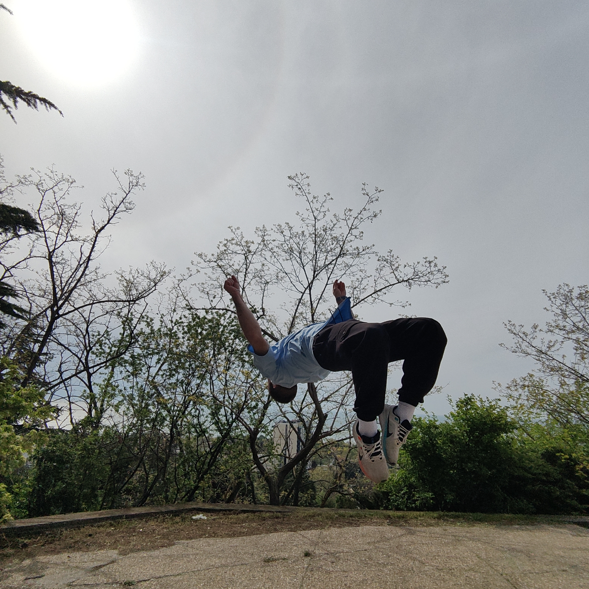 A man is in mid-air, upside down, seemingly performing a backflip. He is wearing a light blue T-shirt, dark-colored pants, white socks, and sneakers. The background shows trees and a cloudy sky with a bright sun, suggesting an outdoor park or green space. A concrete surface or pathway is visible in the foreground. In the background, a tall white building can be seen between the trees.
Based on visual clues, this photo was likely taken in a park in Varna, Bulgaria, with the building potentially being located within the city. The subject appears to be engaged in acrobatics or parkour.