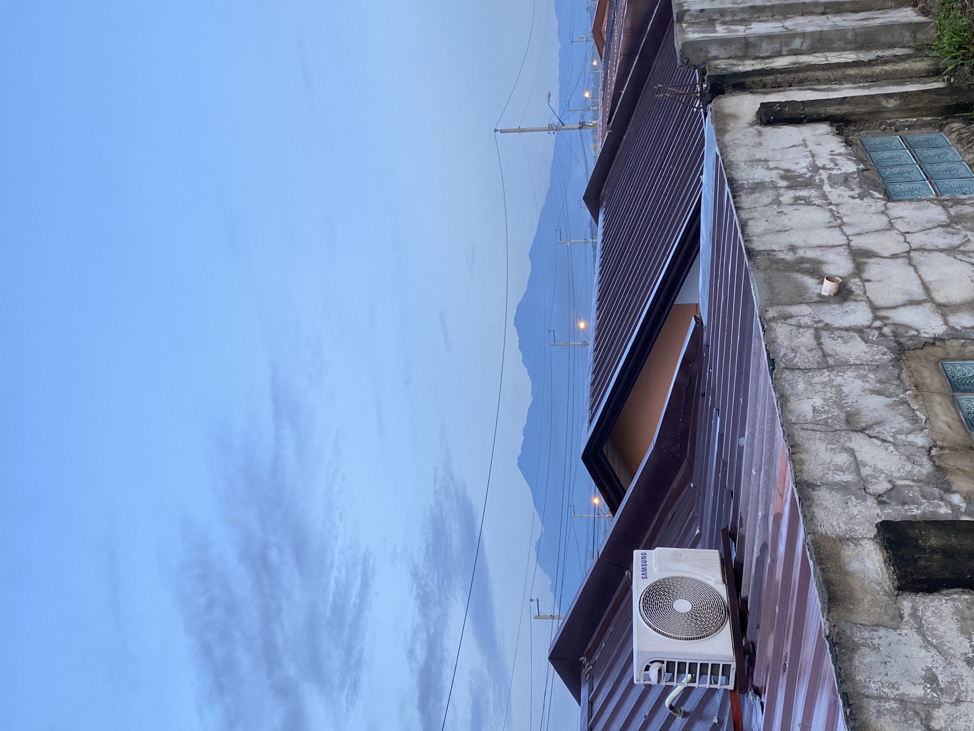 This image, correctly oriented vertically, captures a quiet urban landscape scene, likely at dawn or dusk, in Davao City, Philippines.
In the foreground, a reddish-brown corrugated metal roof is prominent, with a white "SAMSUNG" brand air conditioning unit mounted on it. To the right, a weathered concrete structure, possibly a patio or part of an adjacent building, features embedded square blue glass blocks. This concrete surface shows cracks and wear, and a small, light-colored cylindrical object rests upon it. Concrete steps are partially visible ascending on the far right.
The midground reveals multiple utility poles with a network of power lines stretching across the frame. Several streetlights on these poles are illuminated, emitting a warm yellow glow that contrasts with the cooler tones of the sky. Other corrugated roofs are visible further back, interspersed among the utility infrastructure.
In the background, the silhouettes of distant mountains rise under a light blue, partially cloudy sky. The clouds appear soft and streaky, particularly on the left side of the frame. The subdued lighting, with illuminated streetlights against a non-daylight sky, suggests either early morning or late evening.
No people are visible, and there is no discernible activity taking place. The scene conveys a sense of calm and stillness in an urban setting.