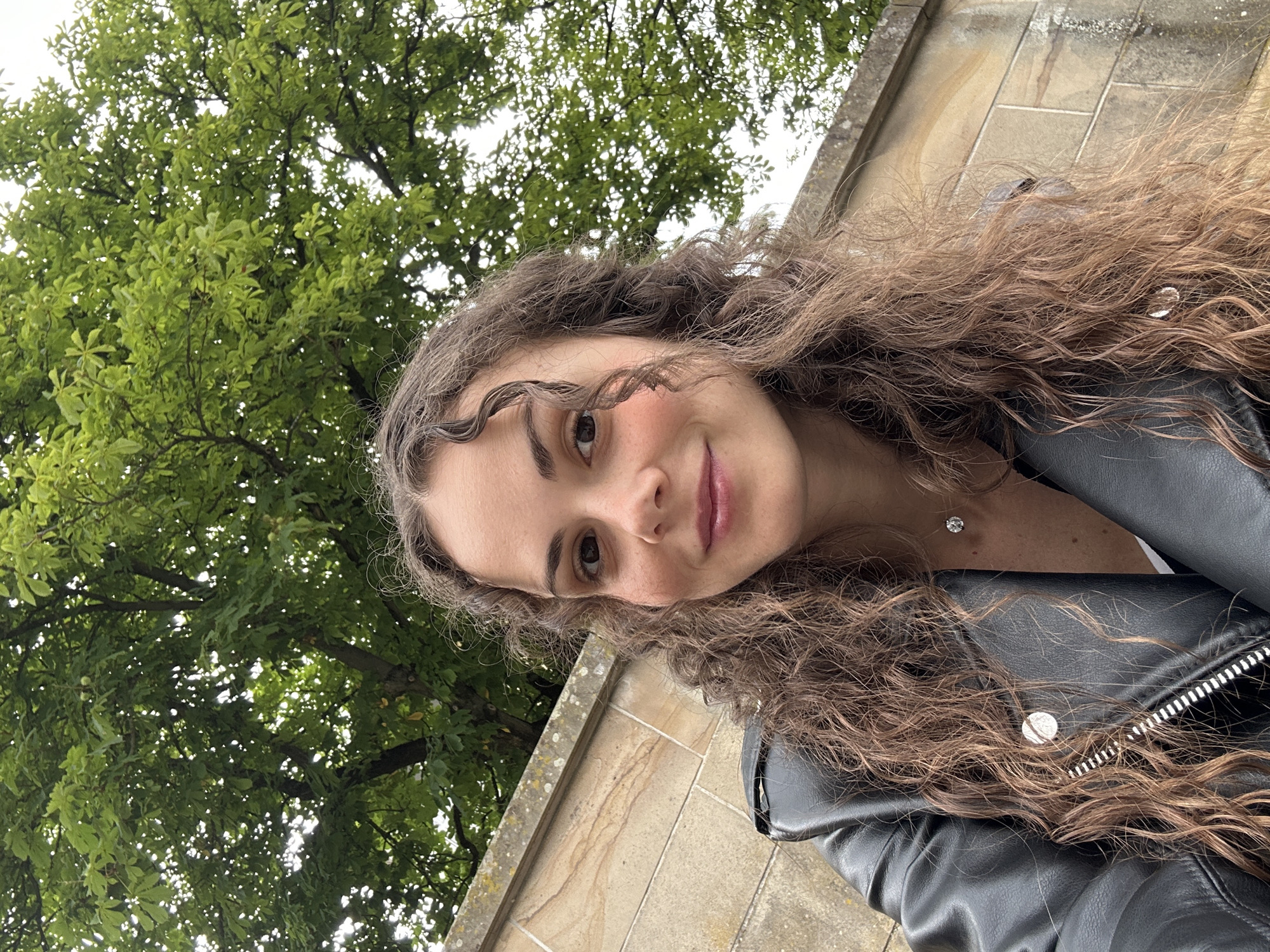 A young woman with long, curly brown hair is captured in a selfie from the chest up, outdoors. She is wearing a black leather jacket with a prominent zipper and a white top underneath, accessorized with a diamond necklace. Her head is tilted slightly as she smiles faintly, looking towards the camera. Behind her, a stone wall forms the lower background, with the dense green foliage of a tree and the overcast sky above. The lighting is soft and natural, suggesting an outdoor setting during the daytime.