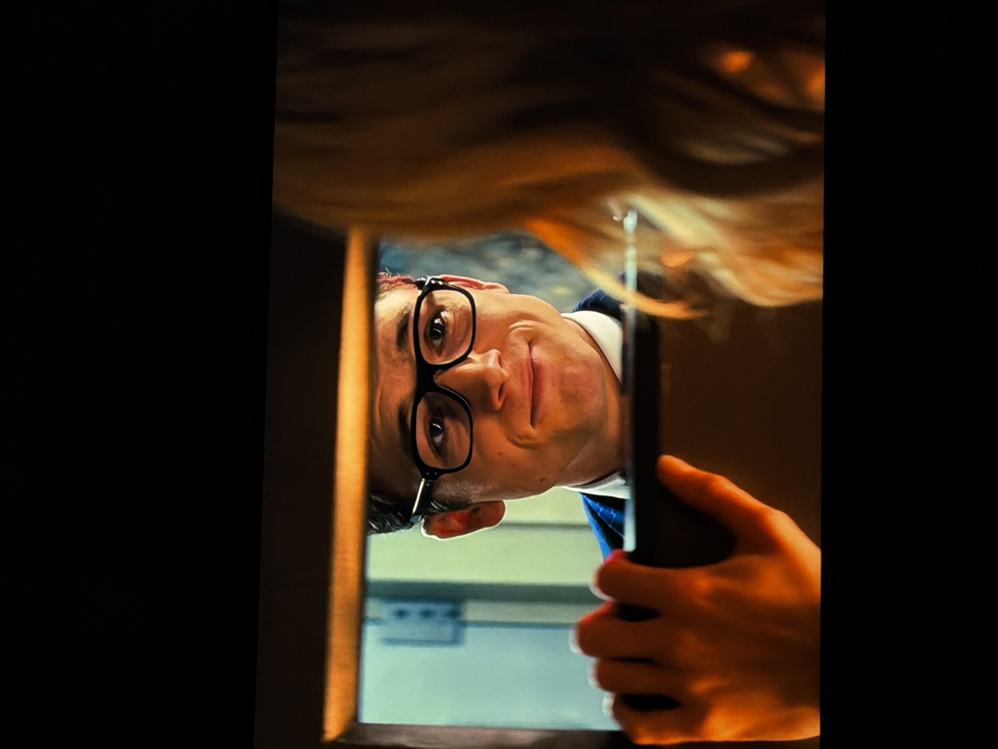 A close-up shot captures a man's face as he peeks through a narrow, dark opening, possibly a gap or a door frame. His head is tilted sharply to the right, and he wears distinctive black-rimmed glasses. A subtle smirk or mischievous smile plays on his lips, and his eyes are directed upwards within the frame, suggesting he is observing something or someone from below. He is dressed in what appears to be a dark suit or jacket with a white collar.
His hand, visible in the lower right, holds a dark, rectangular object, possibly a smartphone or a small device, vertically within the opening. Behind the man, a blurred background of light blue-green suggests an indoor wall or room. At the very top of the frame, out of focus, is a cascade of light brown or blonde hair, hinting at another person's presence above the man, perhaps the one being observed. The unusual perspective and the man's expression create a sense of curiosity, humor, or secretive observation. There is no visible text.