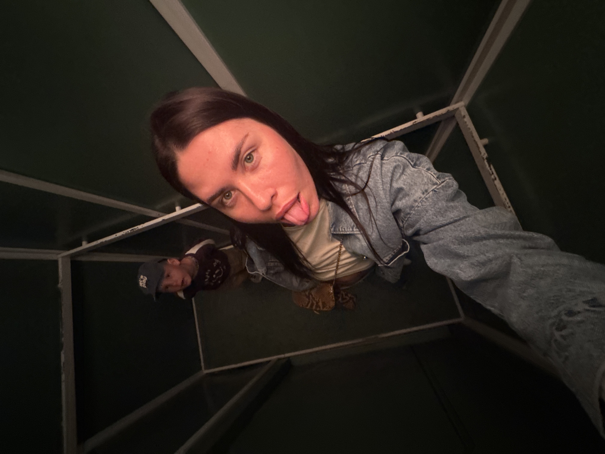 An overhead, high-angle, wide shot captures a young woman and a young boy inside a narrow, dark green, enclosed space with a white frame. The woman, in the foreground, is facing the camera and sticking her tongue out, wearing a denim jacket and a light-colored shirt. She is holding a bag with a tan, patterned strap. The boy, slightly behind and to the left, is looking up with a surprised expression and wearing a dark long-sleeved shirt and a blue baseball cap. The location is identified as Chaikovsky, Russia.