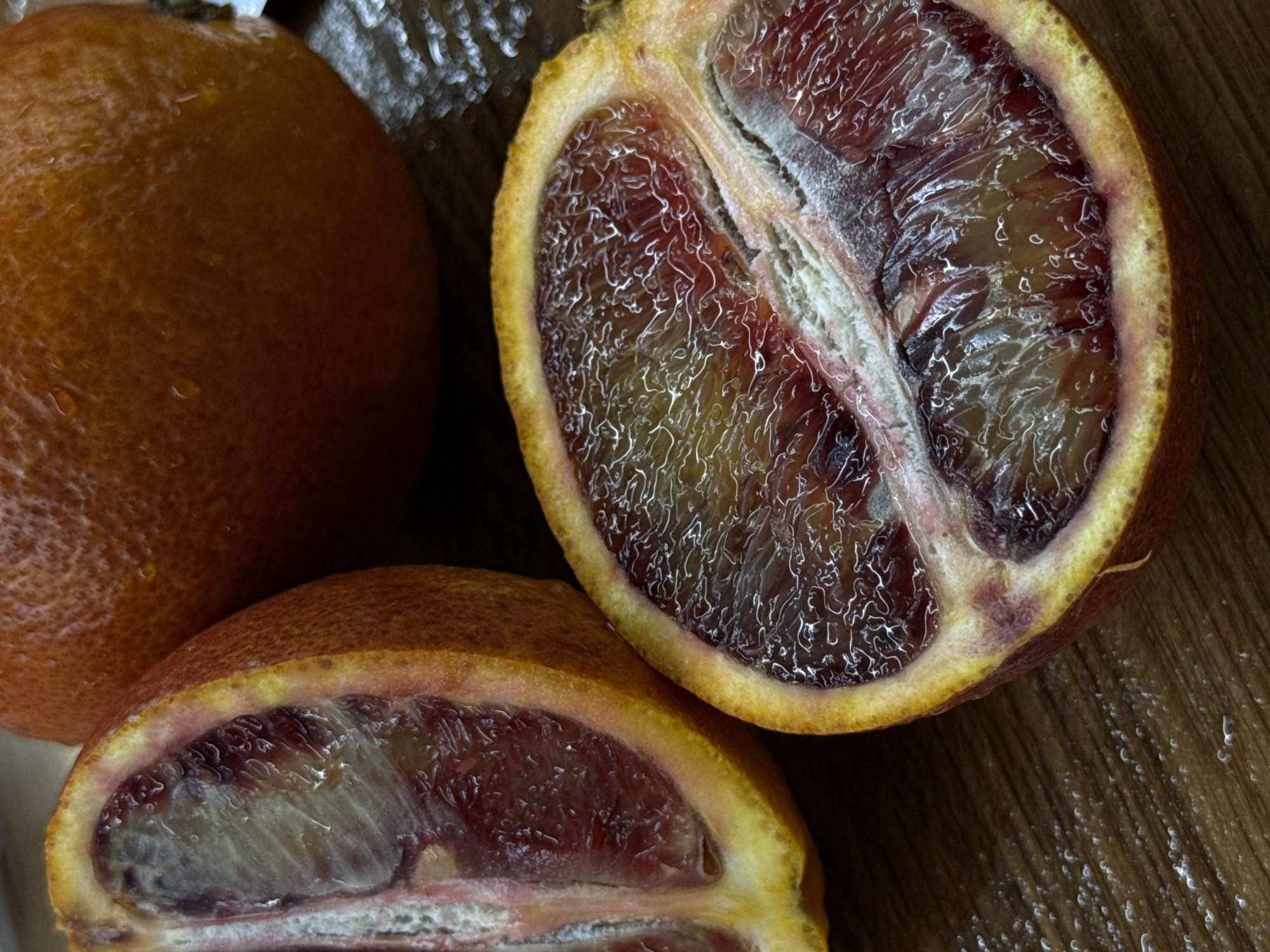 A close-up, dimly lit still life photograph captures three blood oranges, likely on a wooden surface in Pervomaysky, Russia. One whole orange is positioned on the left, displaying its textured, deep orange-red skin, which glistens with a few small water droplets.
To the upper right, a half-cut blood orange reveals its striking interior. The flesh is a rich, dark red, almost purple, with discernible white membranes separating the segments. The juice vesicles within the segments appear somewhat textured or shriveled, creating an intricate, almost lace-like pattern of lighter highlights against the dark pulp. The thick rind transitions from a bright yellow-orange exterior to a reddish hue where it meets the dark fruit.
Another half-cut blood orange is visible in the lower part of the image, partially obscured. Its exposed flesh similarly exhibits the characteristic deep red coloration and white segment divisions, though some areas appear slightly lighter or more translucent.
The background and foreground consist of a wooden surface with a visible grain, which appears somewhat damp or reflective in certain areas, particularly towards the right side of the image. The lighting emphasizes the vibrant, deep colors of the oranges and the intricate details of their cut surfaces. No activities are taking place, and no text is visible in the image. The scene conveys a simple, natural arrangement of fruit.