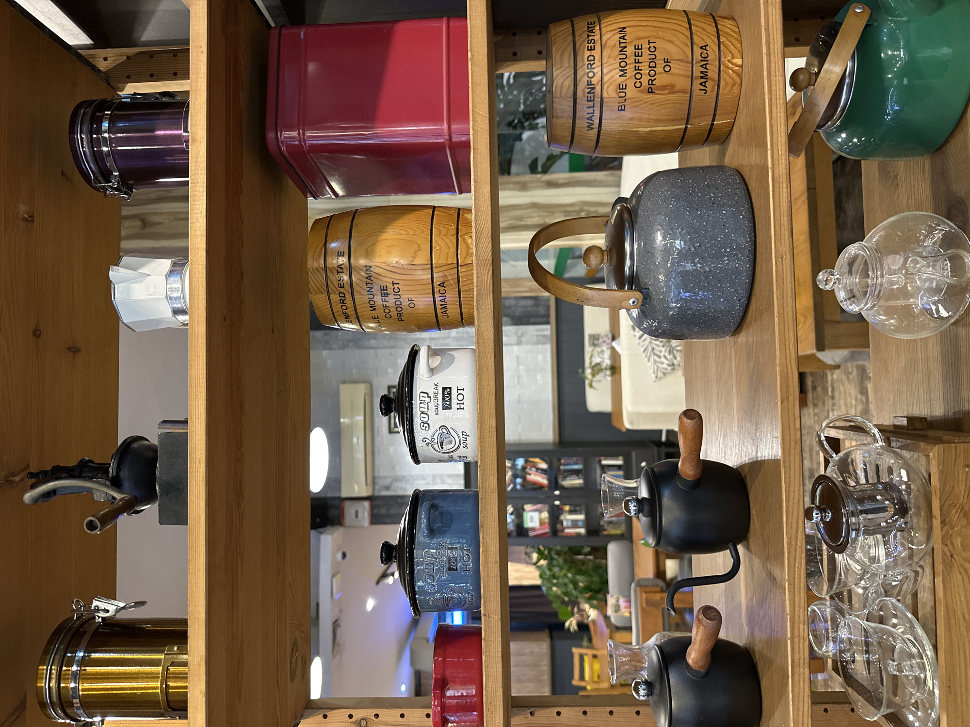 The image shows several shelves in what appears to be a cafe or shop in Kuta Selatan, Indonesia. The shelves are made of light-colored wood and are filled with various kitchenware and decorative items.
On the top shelf, from left to right, are a tall gold canister, a black coffee grinder with a spout and handle, a white moka pot, and a dark purple canister.
The second shelf from the top contains a red bowl on the far left, a blue soup bowl with "SOUP BREAK 100% HOT" written on it, a white soup bowl with similar text, a wooden barrel labeled "ENFORD ESTATE BLUE MOUNTAIN COFFEE PRODUCT OF JAMAICA," and a tall red rectangular tin.
The third shelf features a dark gray kettle with a wooden handle on the left, a black kettle with a long spout and wooden handle, and another wooden barrel labeled "WALLENFORD ESTATE BLUE MOUNTAIN COFFEE PRODUCT OF JAMAICA" on the right.
The bottom shelf displays a wooden tray with several clear glass teacups and saucers, a clear glass teapot with a metal infuser, a small clear glass teapot, and a green kettle with a wooden lid handle.
In the background, through an open space between shelves, there are tables and chairs, suggesting a seating area. A bookshelf filled with books is also visible in the background. The lighting appears to be artificial, with some ceiling lights visible. No people are present in the image.