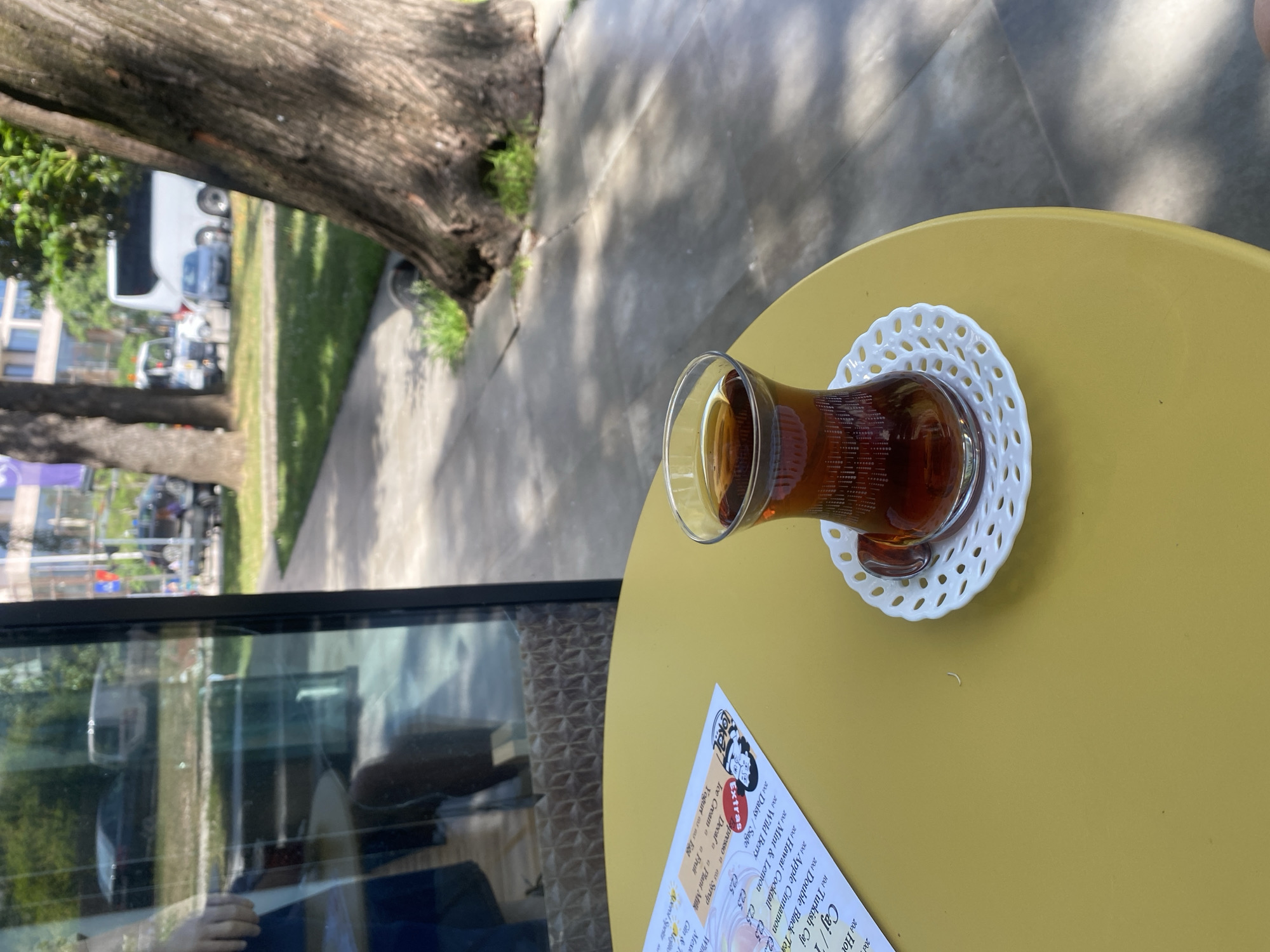 On a yellow table, outdoors in Budva, Montenegro, sits a glass of tea on a white, patterned saucer. A menu is partially visible at the lower left. The setting includes a view of a tree, a sidewalk, and street activity seen through a window or reflective surface.