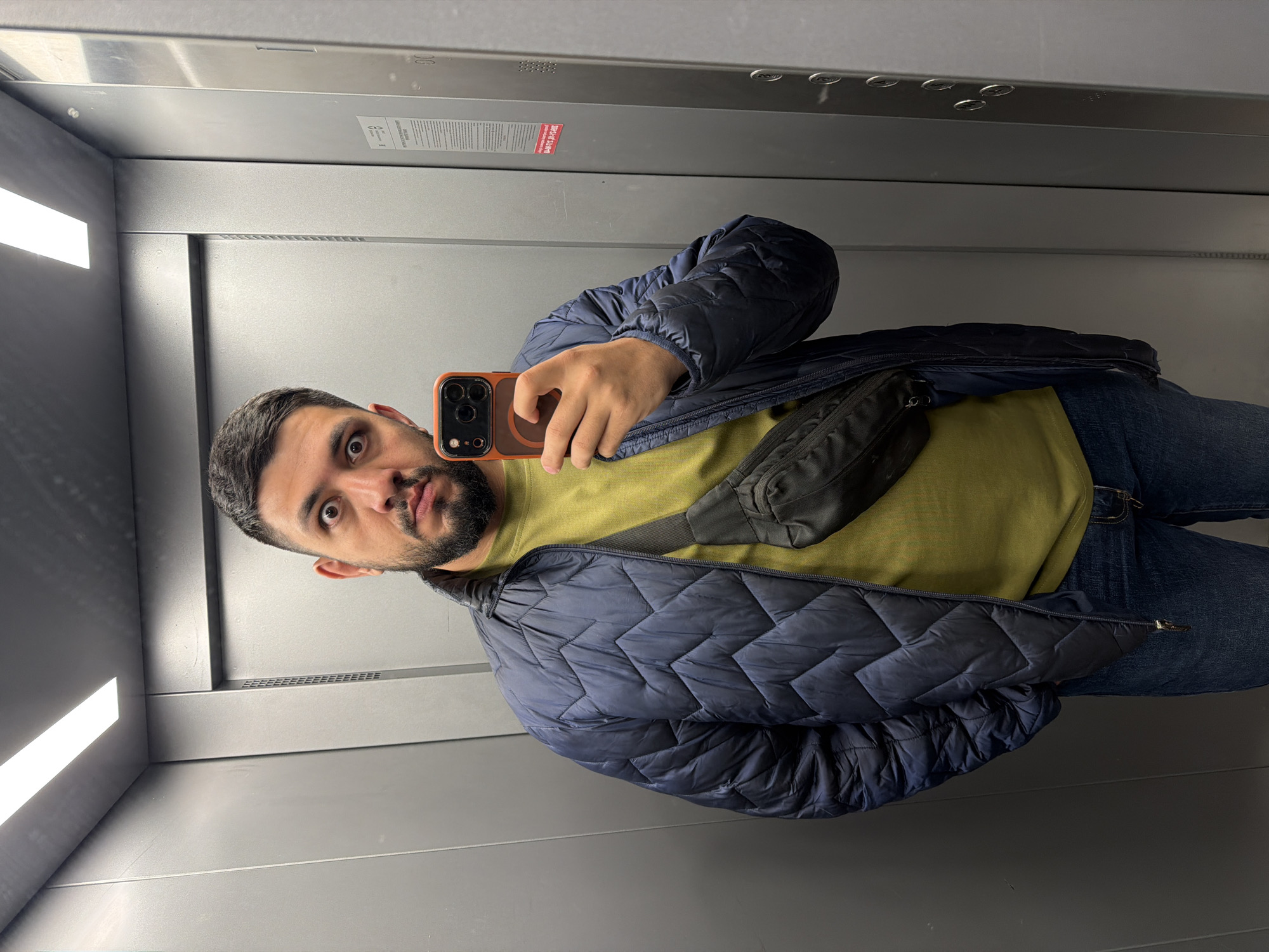 A man with dark hair and a beard is taking a selfie in what appears to be an elevator. He is wearing a blue quilted jacket over a mustard-yellow top and blue jeans. A black fanny pack is slung across his body. The elevator interior is gray, with bright rectangular lighting on the left. There is signage visible above him. There's no information to indicate the image was taken in the city of Krasnoyarsk, Russia.