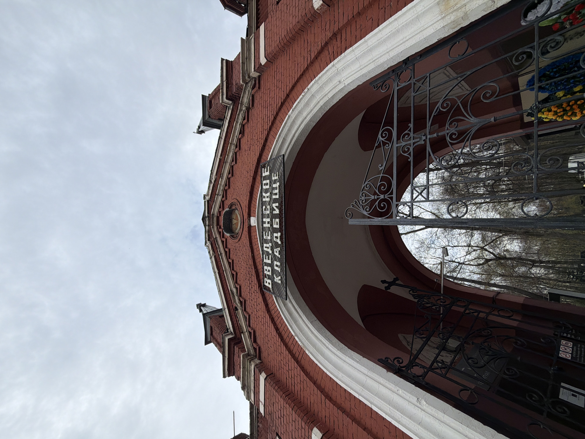 A low-angle shot captures the grand, arched entrance of a red brick building, identified as the Vvedenskoye Cemetery in Moscow, Russia. The perspective looks upwards towards an overcast sky, with the building's facade dominating the frame.
The building is constructed from dark red brick, featuring a large, symmetrical archway outlined with contrasting white trim. Decorative elements and a tiered roofline are visible at the top, hinting at intricate architecture. Hanging vertically within the arch is a black sign with white Cyrillic text, which reads "ВВЕДЕНСКОЕ КЛАДБИЩЕ," translating to "Vvedenskoye Cemetery."
The archway is secured by an ornate, black wrought-iron gate with intricate scrollwork and patterns. Through the gate, the bare branches of trees can be seen against the muted, cloudy sky, suggesting late autumn or winter. Inside the arch, to the right, vibrant artificial flowers or memorial wreaths in yellow, blue, and other colors are visible, commonly used at cemeteries. A security camera is also discernible within the entrance.
The scene conveys a sense of solemnity and historical significance, typical of a major cemetery entrance. The overcast weather contributes to a subdued atmosphere. No people or specific activities are present, focusing solely on the architectural gateway.