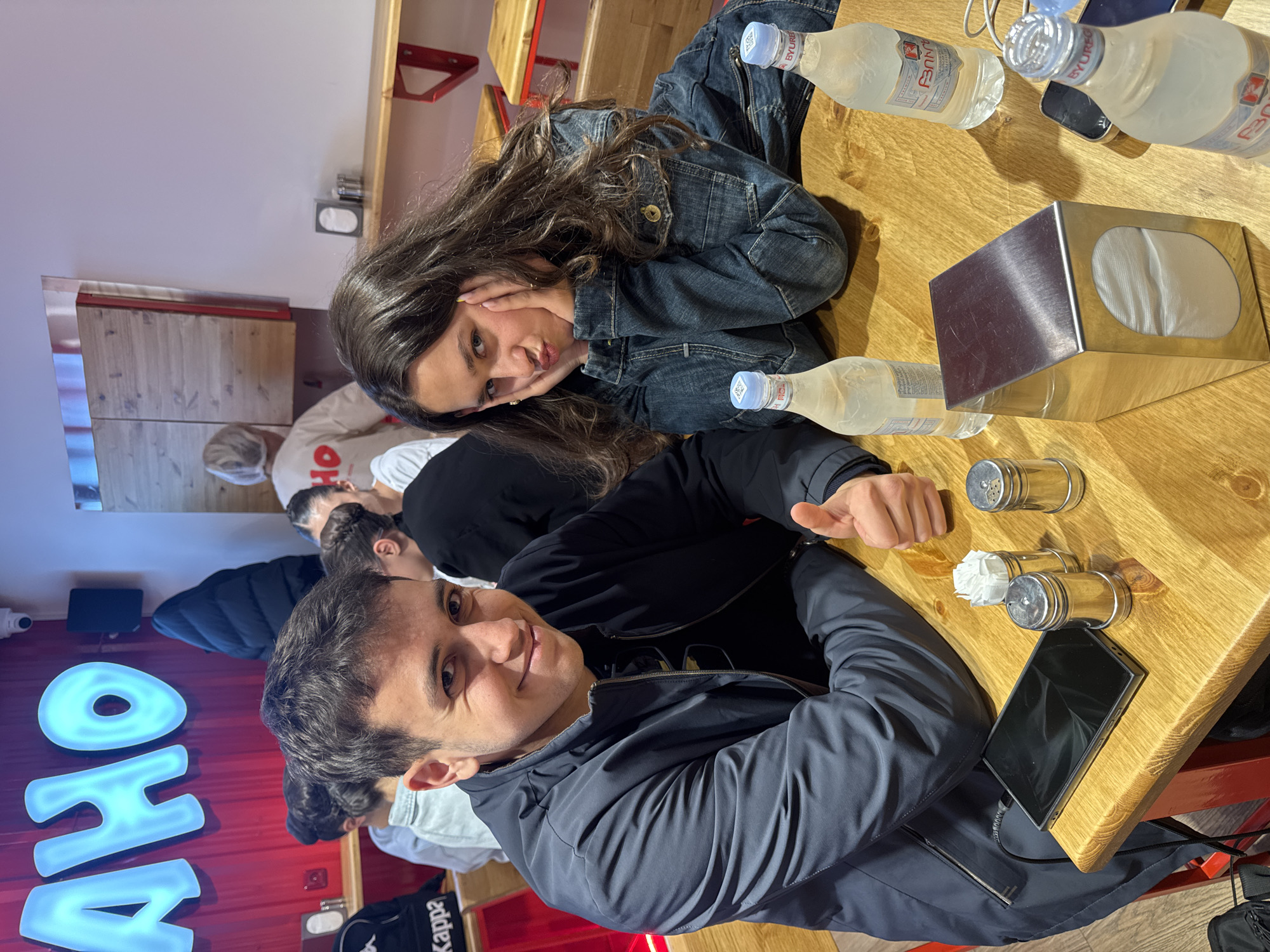 The original image, rotated 90 degrees counter-clockwise, captures an indoor scene within a casual cafe or restaurant setting, likely in Yerevan, Armenia.
Two young individuals are prominently seated at a light wooden table. A young woman with long, dark, wavy hair, wearing a denim jacket, looks towards the camera with a playful pout, resting her chin on her hands. Beside her, a young man with short, dark hair, dressed in a dark blue or gray jacket, smiles brightly at the camera and gives a thumbs-up gesture. Both appear relaxed and happy.
The table holds several items: multiple clear plastic bottles of water with labels showing "BYUREB" and other text (likely Byuregh mineral water, common in Armenia), a metallic napkin dispenser with white napkins, two metallic salt and pepper shakers, and a black smartphone connected to a charger.
In the background, other patrons are partially visible, including one person wearing a light-colored top with the letters "HO" on it. A large, illuminated blue sign spelling "OHA" (or similar, partially obscured) is mounted on a red wall, and a security camera is also visible. Red structural elements, possibly chair or table frames, are present throughout the background. A mirror on the left side of the frame reflects parts of the interior. The atmosphere suggests a casual social gathering or meal.