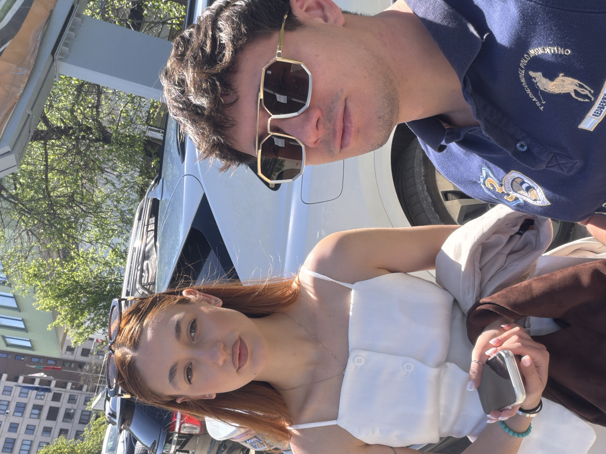 Two individuals pose for a selfie outdoors. The man, wearing sunglasses and a blue polo shirt with an emblem, is positioned above and to the right of the woman. The woman, with auburn hair and a white top, looks directly at the camera and holds a phone in her hands. They are standing in front of a white car. The surrounding environment includes urban elements like buildings and trees, suggesting a city setting, likely Sofia, Bulgaria.