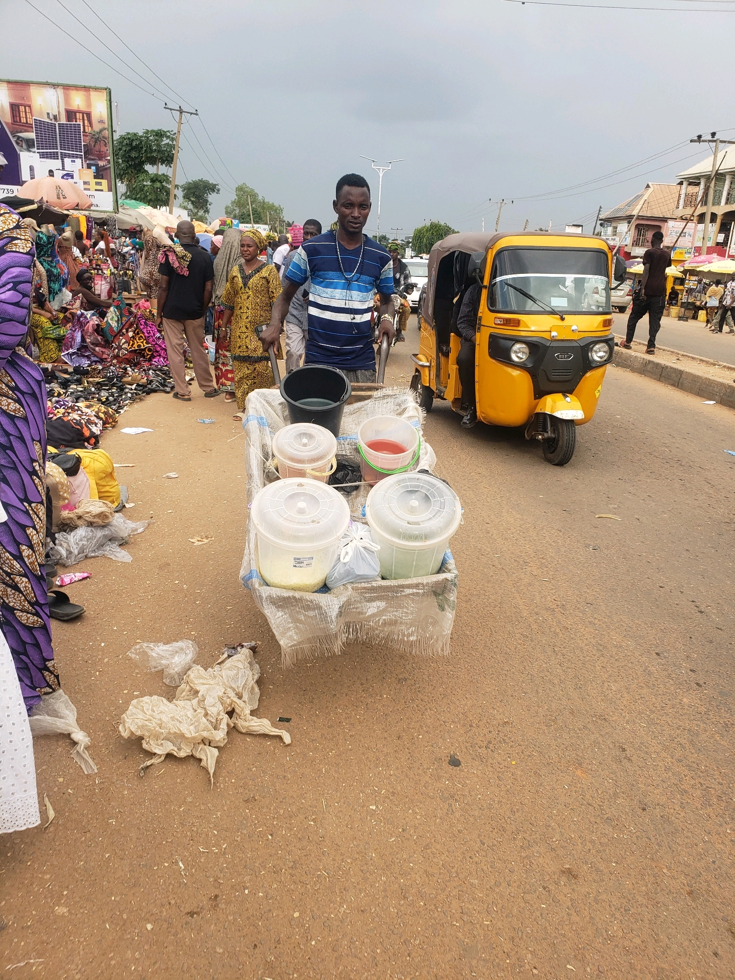 In Sabon Tasha, Nigeria, an outdoor marketplace is captured on an overcast day. A man, wearing a blue and white striped shirt, stands in the foreground, pushing a cart loaded with buckets and containers. The cart's contents include a black bucket, a clear bucket with red liquid, and a large white bucket filled with a yellowish substance. The cart itself is covered in a clear plastic sheeting. Behind the man, a yellow auto-rickshaw, commonly known as a "keke," is parked on the side of the road. The marketplace is bustling with activity, featuring stalls with colorful fabrics and various goods. People are seen browsing and interacting, with many women dressed in traditional Nigerian attire. The road is paved, and the surrounding environment suggests a busy urban setting with buildings and utility poles visible in the background. Scattered plastic waste litters the ground, indicating a less-than-pristine urban environment.