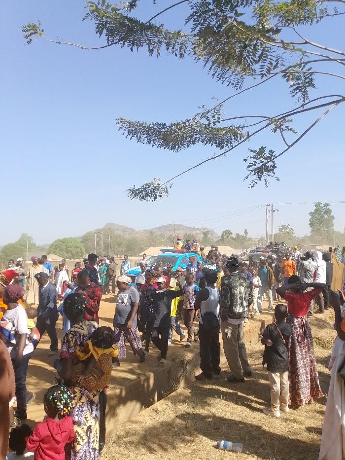The scene, located in Kpak, Nigeria, depicts an outdoor gathering under a clear, bright blue sky with partial obstruction from leafy tree branches in the upper frame. The ground is dry and dusty, featuring a shallow, excavated trench running horizontally through the mid-ground. A substantial crowd of individuals, comprising men, women, and children of various ages, occupies the central and lower portions of the image. They are attired in diverse casual and traditional clothing, with some women carrying infants on their backs. The predominant activity involves walking and standing, with individuals generally oriented towards the middle distance. Two light-blue pickup trucks, equipped with roof-mounted lights and personnel, are visible amidst the crowd in the mid-background. Distant hills, scattered vegetation, and utility poles with overhead lines are present in the hazy background. A single, clear plastic bottle lies discarded on the dry ground in the lower-right foreground.